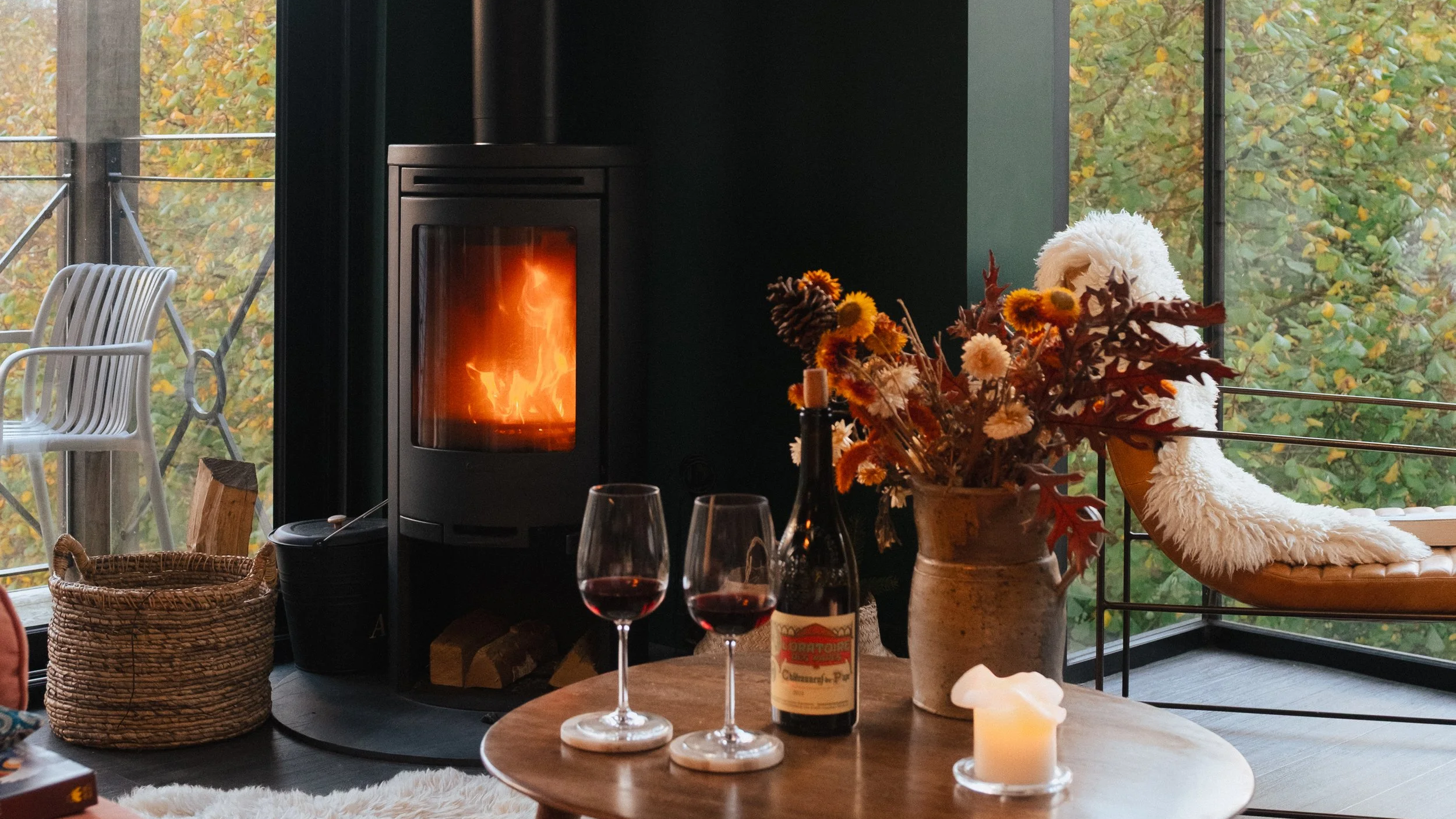 Cozy living room with a lit fireplace, a round wooden table with two glasses of red wine, a bottle of wine, a candle, and a vase with autumn flowers. A plush white blanket is draped over a round lounge chair near large windows showing fall foliage ou