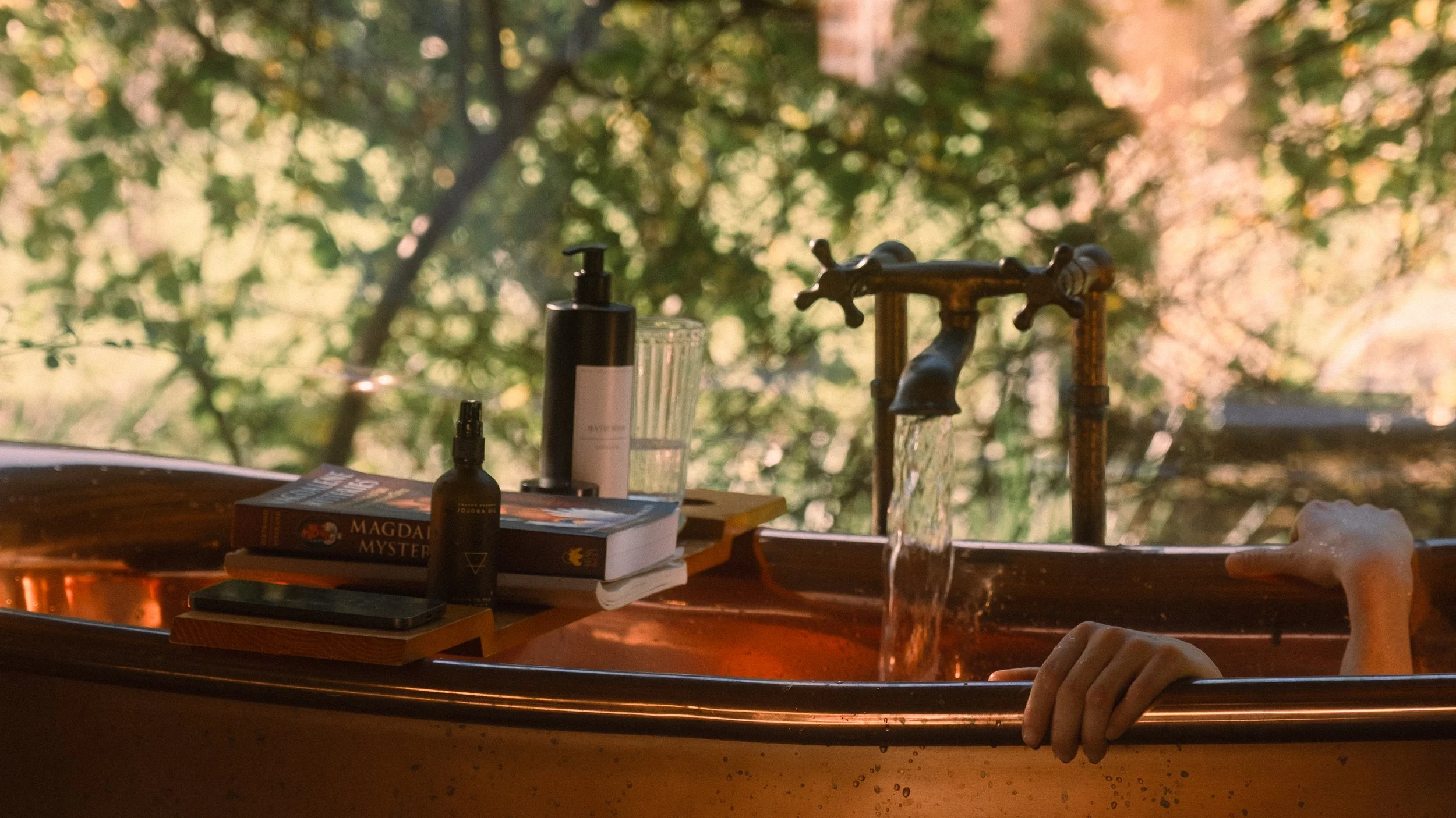 A copper bathtub outdoors with a person’s arm and hand immersed in water, surrounded by trees with green and orange leaves, and various bath items on the edge including books, bottles, and a glass.
