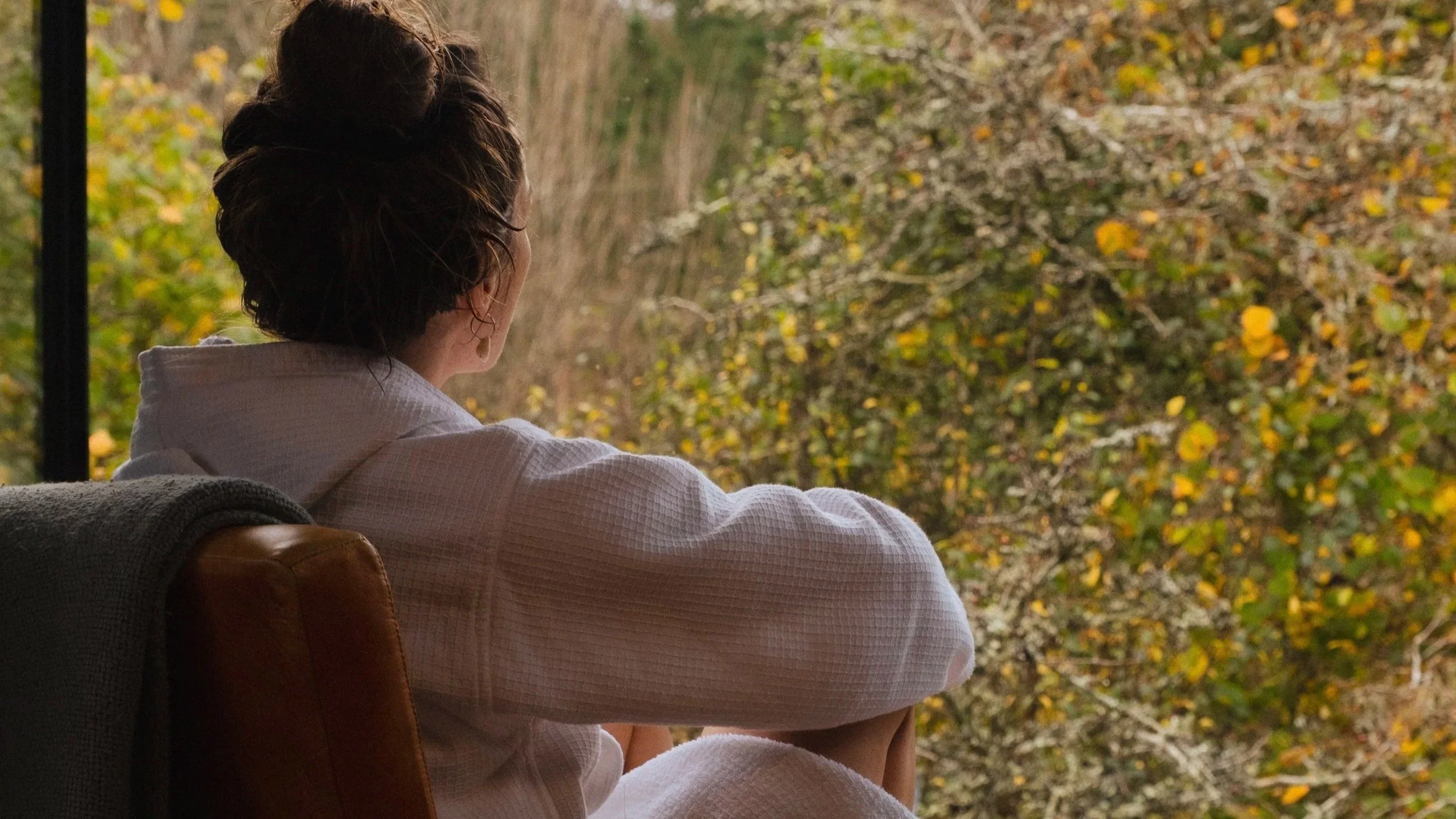 A woman with dark hair in a bun sitting on an armchair, gazing out a window at autumn foliage outside.