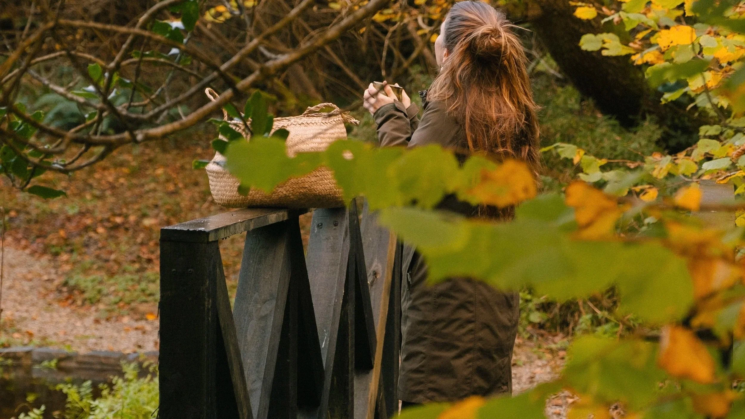 A woman with long brown hair tied in a ponytail, wearing a dark jacket and brown skirt, is sitting on a wooden bridge in a park with fall foliage, holding a small object in her hands. A woven bag is placed beside her on the bridge, and leaves and tre