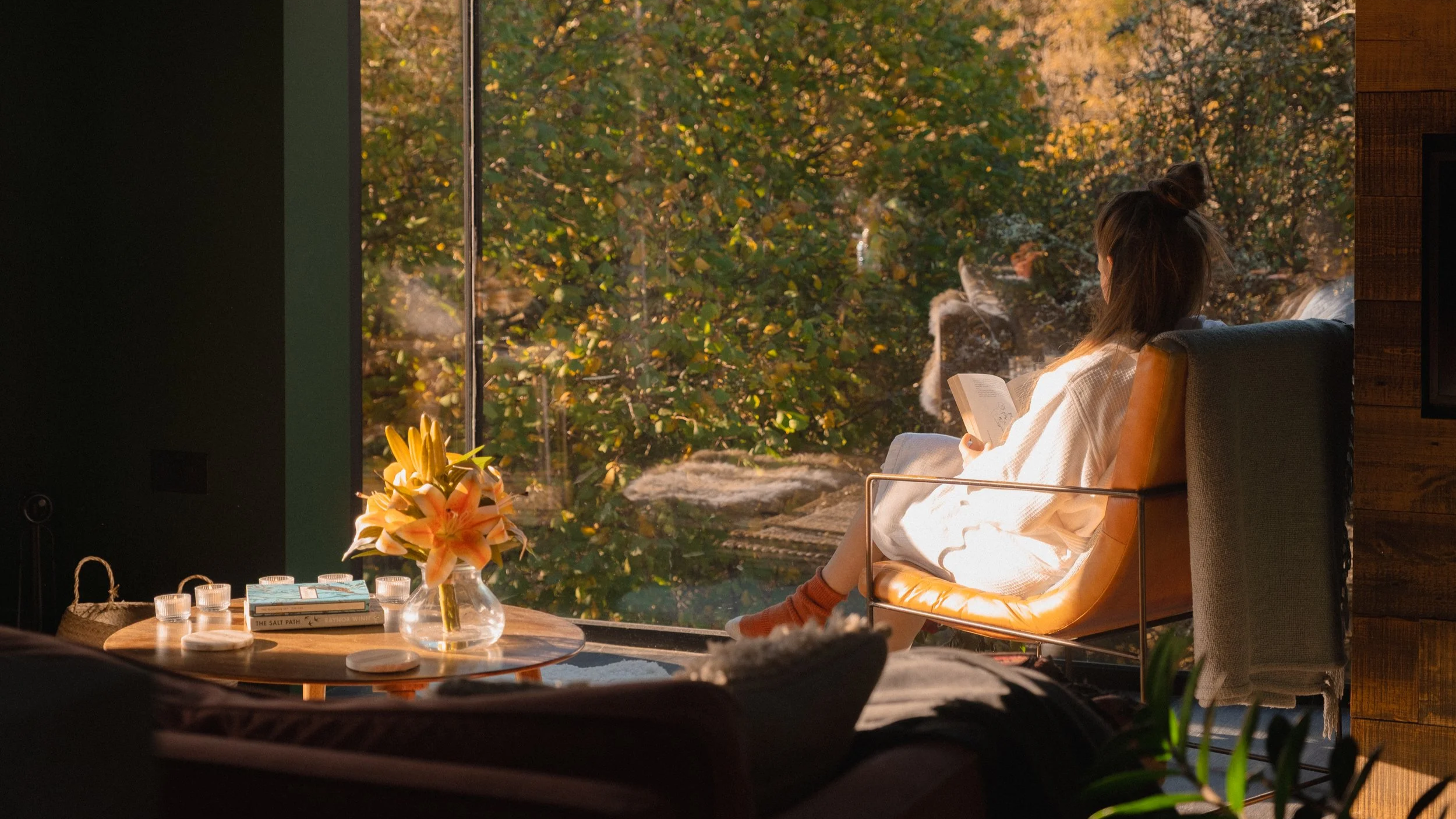 A woman sitting in an orange chair near a large window, reading a book. Sunlight illuminates her and the room, with a view of trees with autumn leaves outside. A round table with books, a bouquet of lilies, and candles is in the foreground.