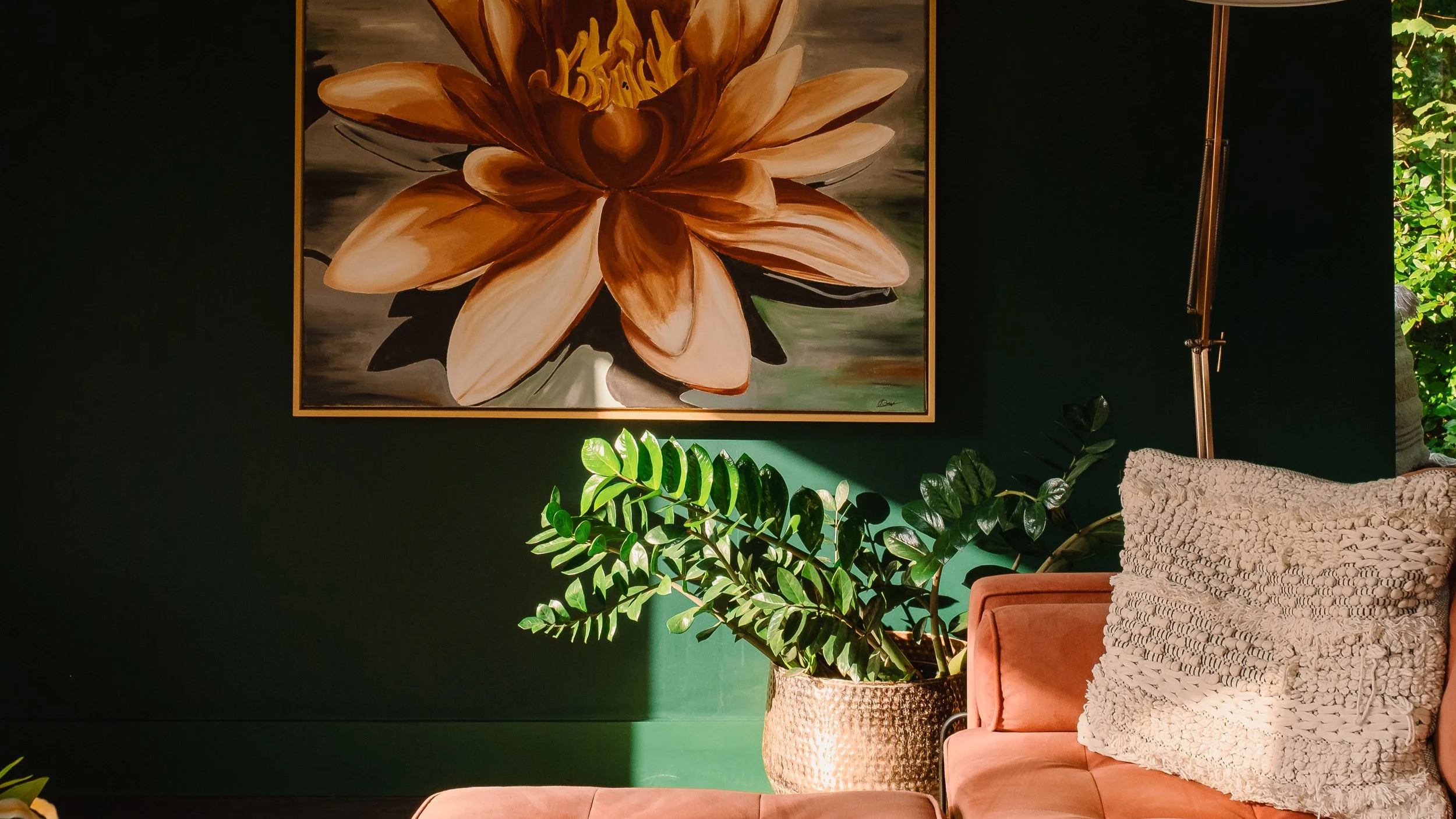 Interior of a living room with a dark green wall, a large flower painting hanging on the wall, a potted plant with glossy leaves, a pink couch with a textured white pillow, and a standing lamp, with sunlight casting shadows.