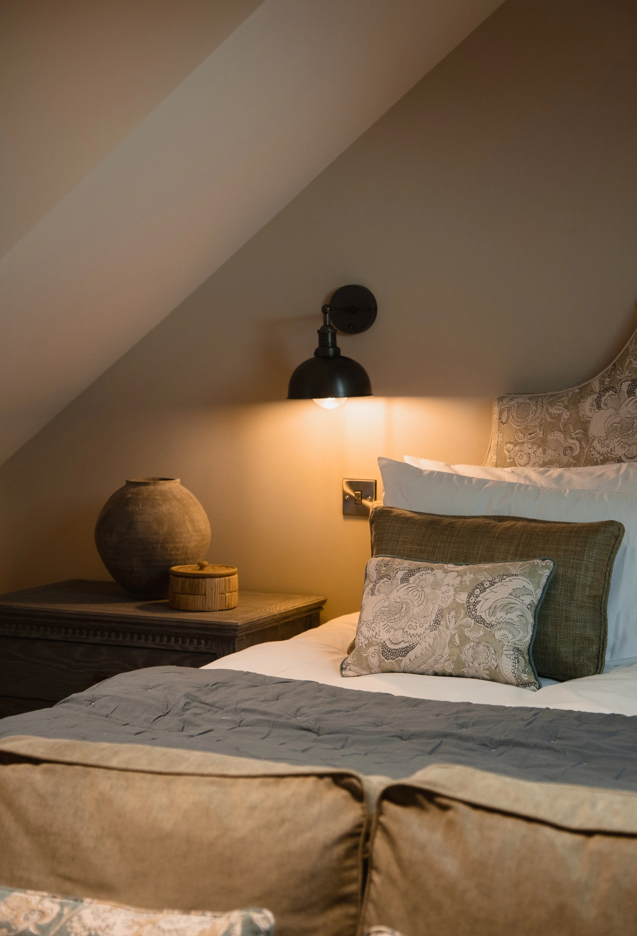 A cozy bedroom with a sloped ceiling, featuring a bed with decorative pillows, a black wall sconce light, and a bedside table with pottery and a small wooden box.