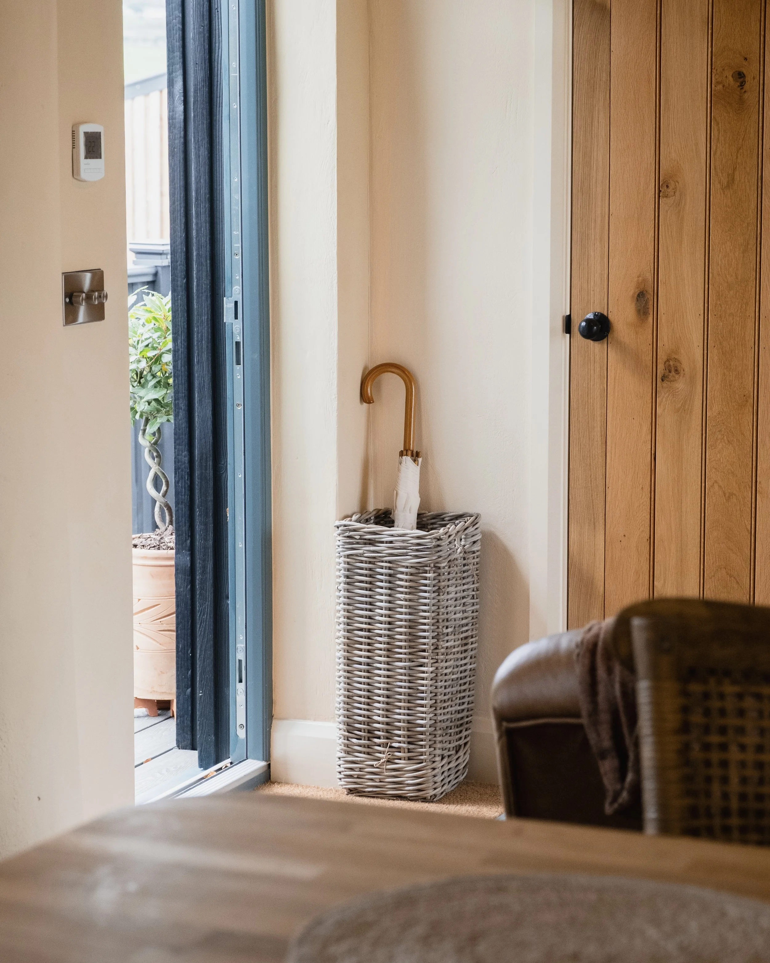 Indoor view of a corner near a porch door, with a wicker umbrella stand holding an umbrella. Part of a table and a chair with a cloth are visible in the foreground.
