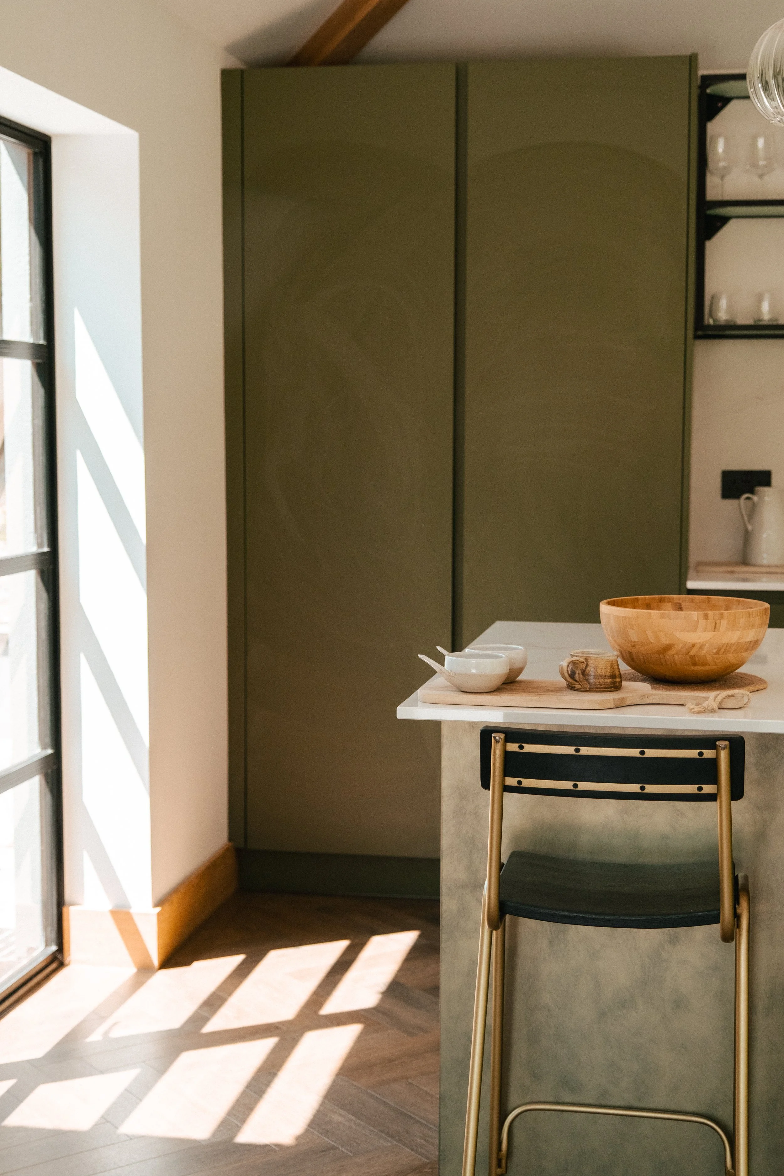 A kitchen corner with a white countertop, a large wooden bowl, two small ceramic bowls, and a small mug placed on a wooden tray, with sunlight casting shadows on the wooden floor.