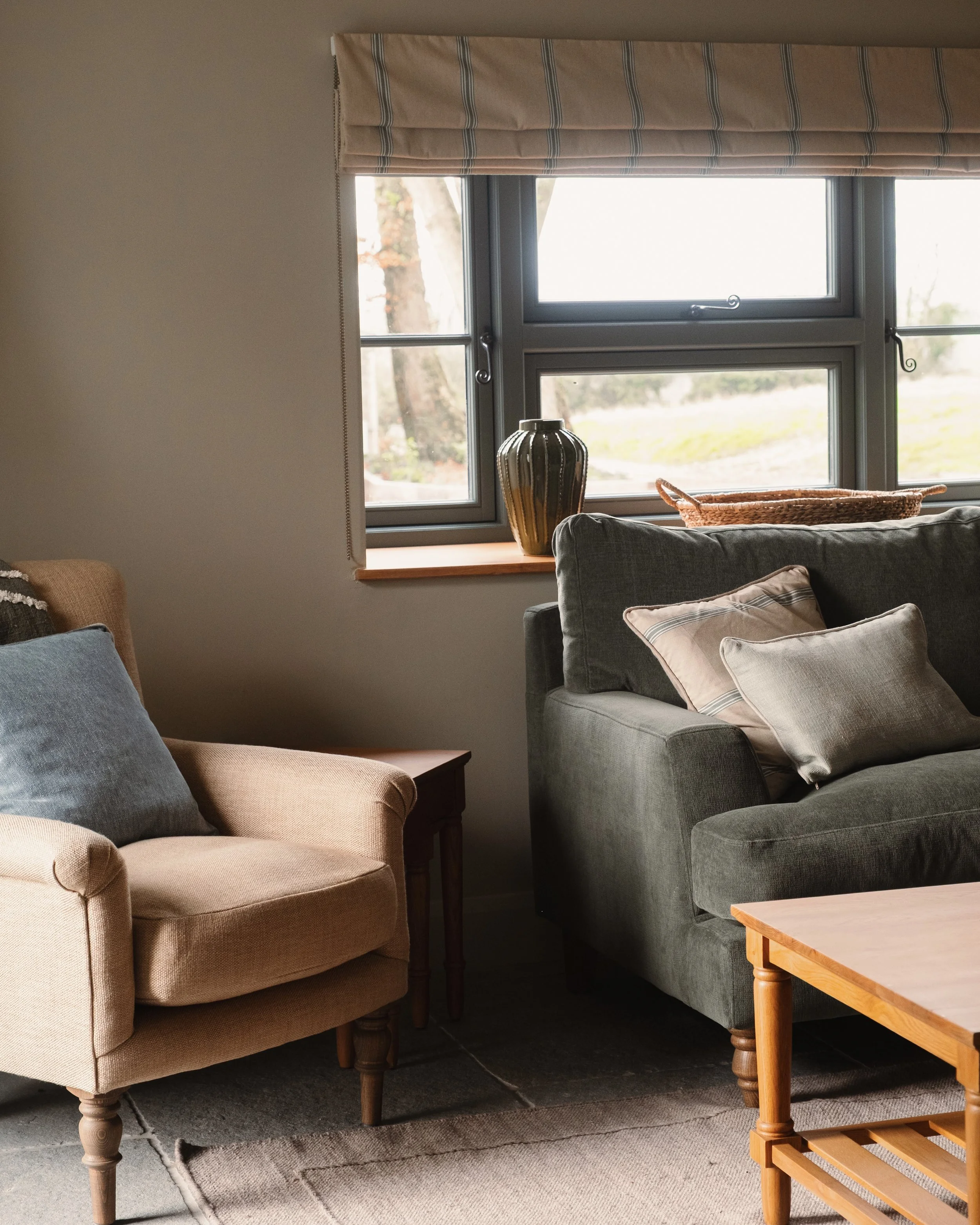 Living room with a beige armchair and a grey sofa, a window with a beige and blue striped curtain, a decorative vase and wicker basket on the windowsill, and a wooden coffee table.