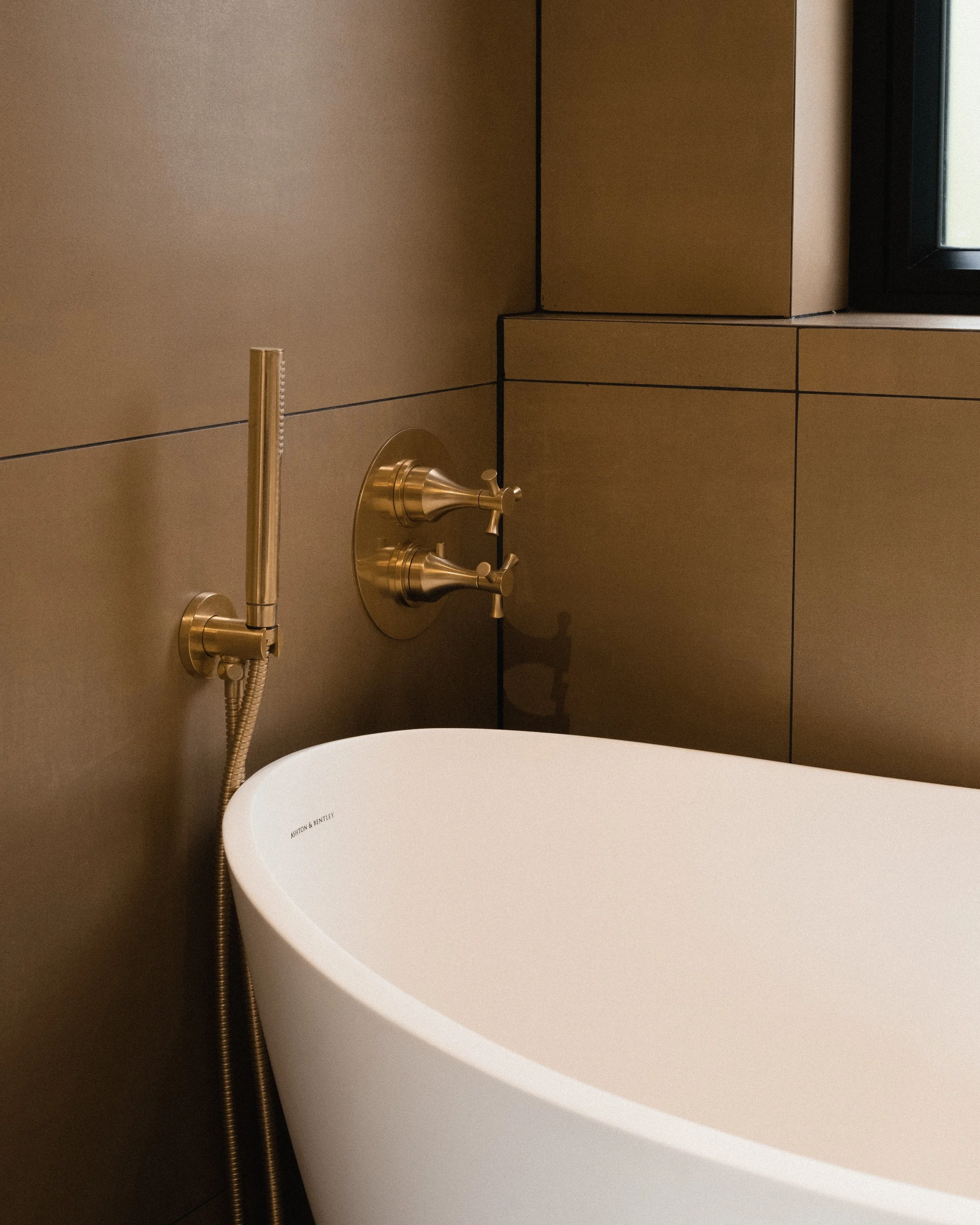 A modern bathroom with a white oval bathtub, bronze-colored wall-mounted faucet with separate handles, a handheld shower spray, and a window with a black frame letting in natural light.