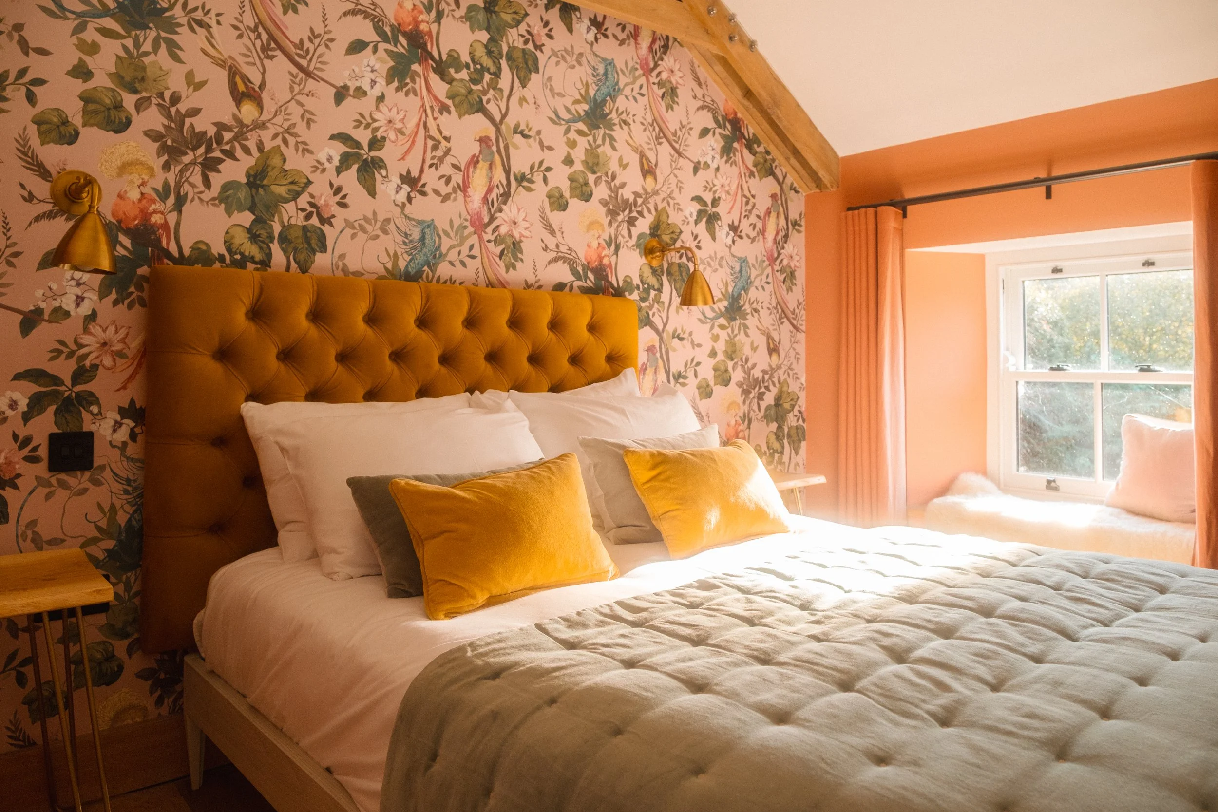 A cozy bedroom with a large bed featuring a mustard-colored tufted headboard, decorated with white and sage green pillows, and a beige quilted bedspread. The wall behind the bed has floral wallpaper with birds and foliage. There are gold wall-mounted