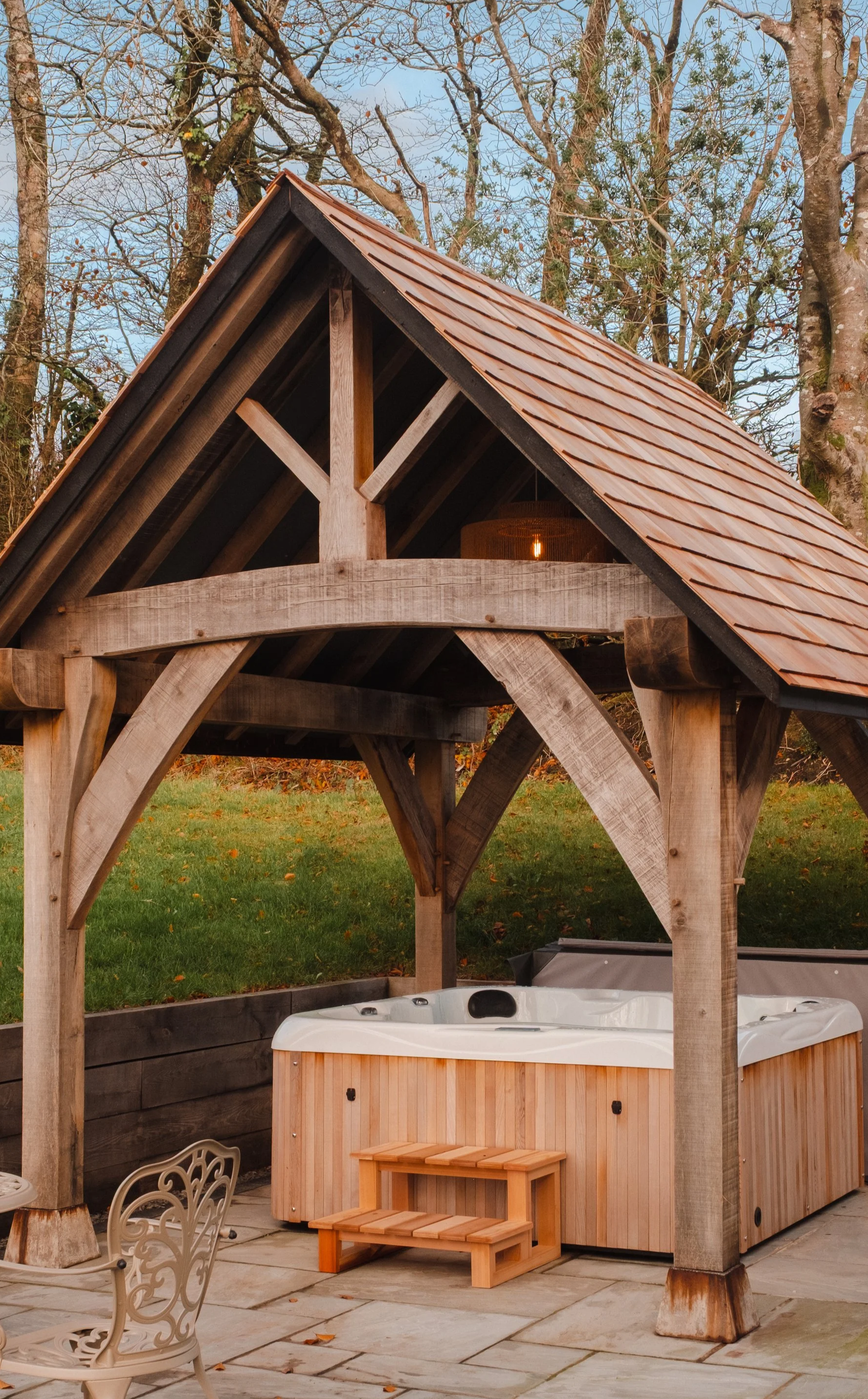 An outdoor wooden hot tub under a wooden pavilion with a shingled roof, set on a stone patio. There are trees and grass in the background.