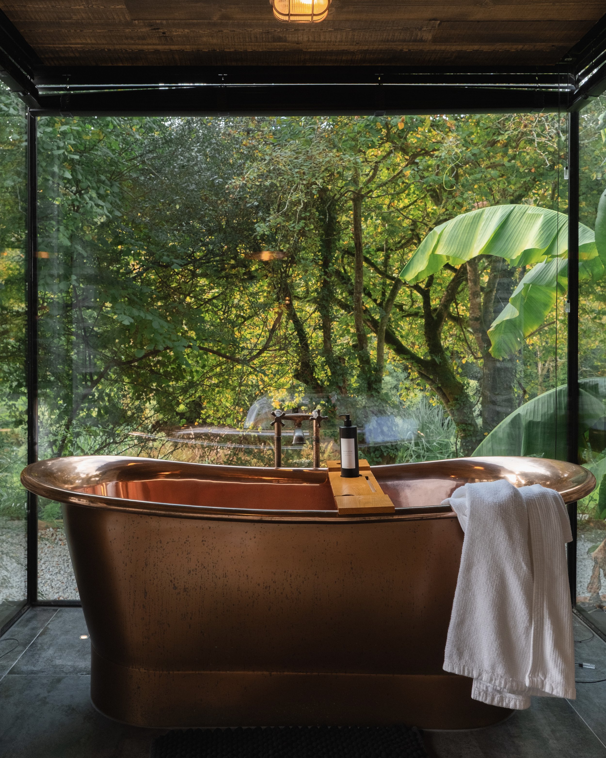 A copper bathtub positioned by a large glass window overlooking lush green trees and plants outside, with a white towel hanging over the edge and bath products on a small wooden tray.