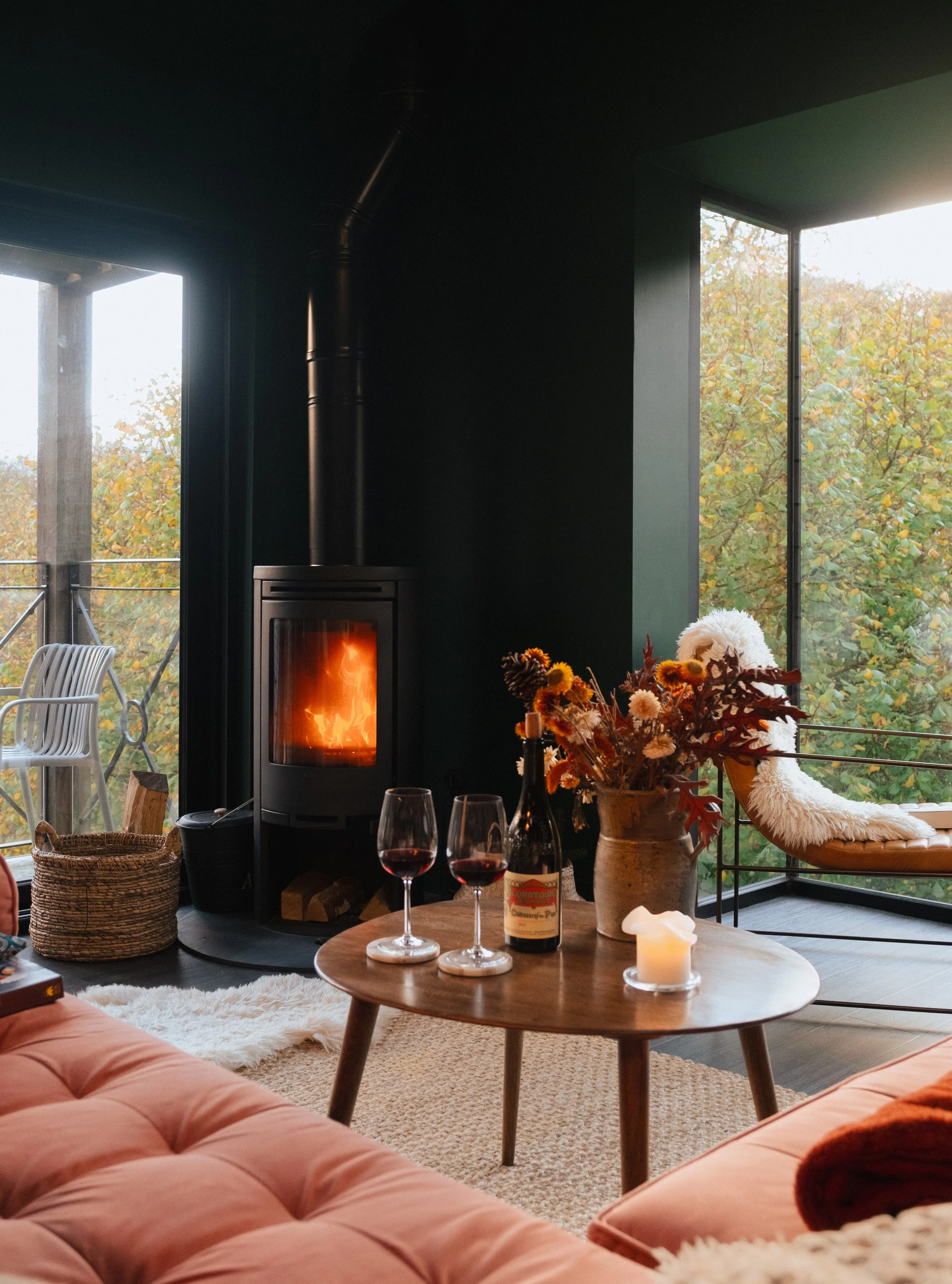 A cozy living room with a lit fireplace, a pink tufted sofa, a wooden coffee table with two glasses of red wine, a bottle of wine, a candle, a vase of autumn flowers, and large windows overlooking a tree-filled balcony.