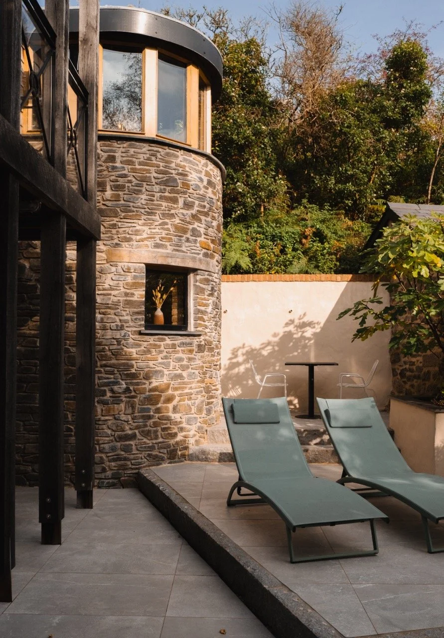 Two lounge chairs with cushions in an outdoor patio area, next to a stone building with large windows and surrounded by greenery and trees.