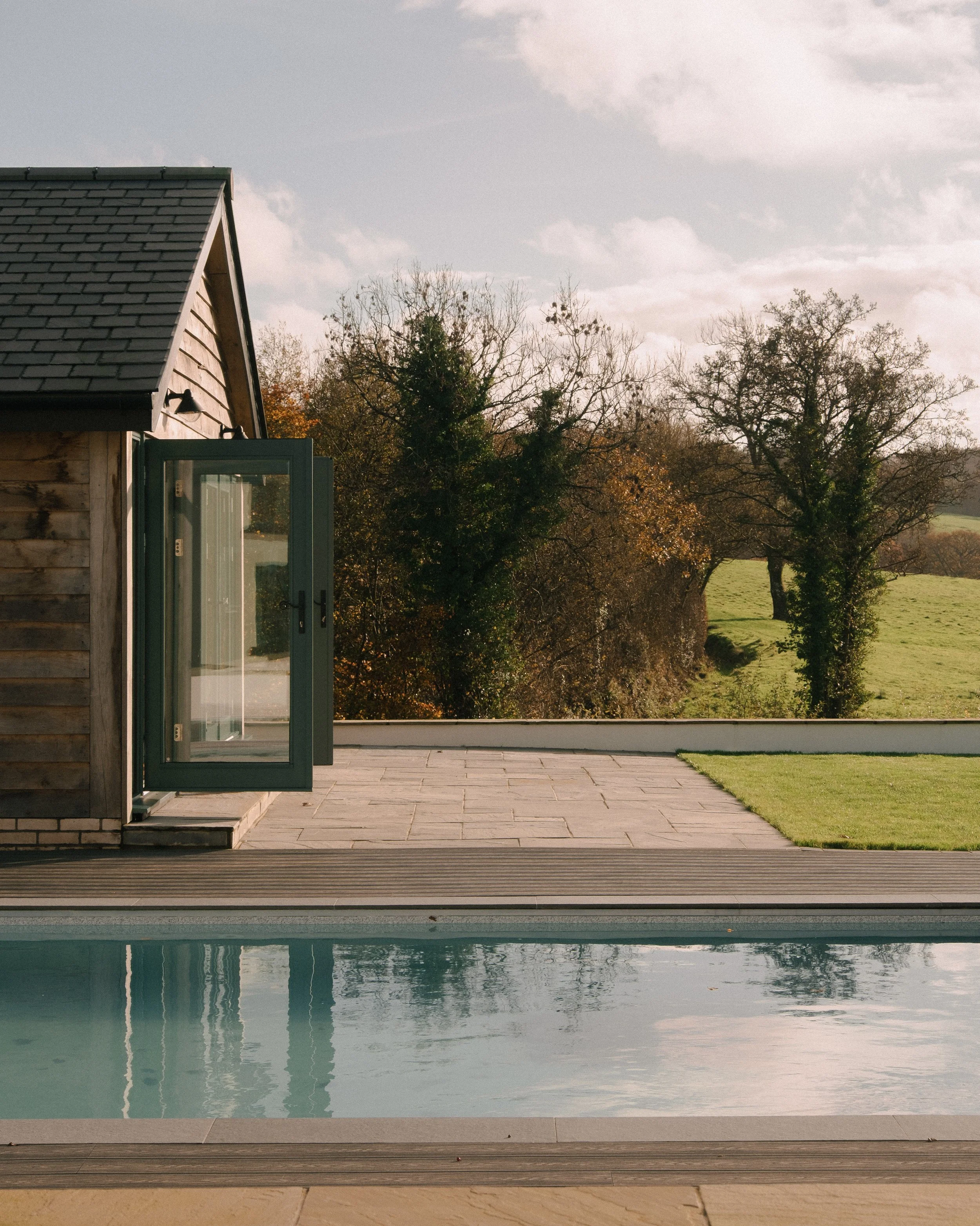 View of a wooden house with an open door leading to a patio and swimming pool, overlooking a grassy landscape with trees and a cloudy sky.