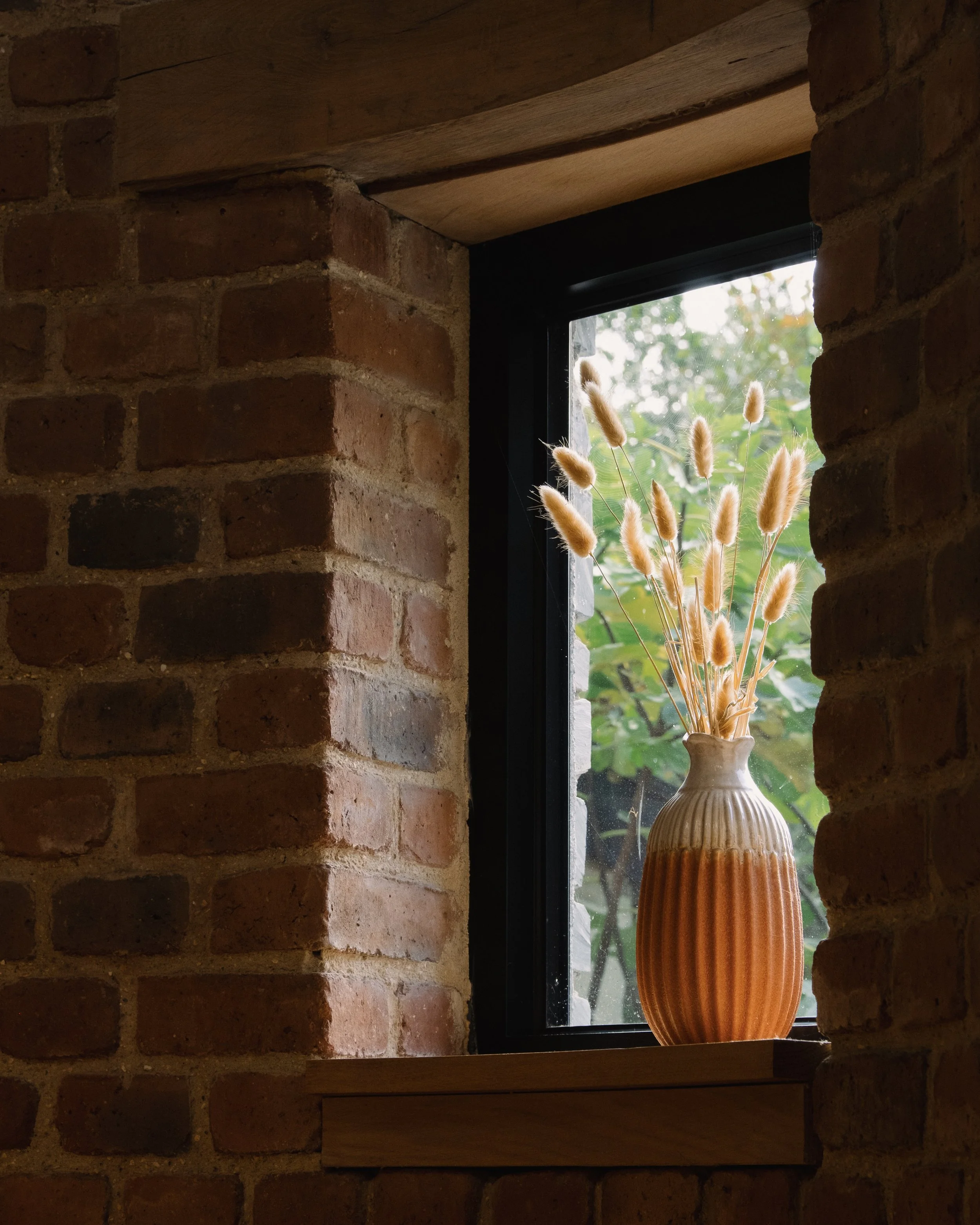 A ceramic vase with vertical ridges and a two-tone glaze, containing dried bunny tail grasses, on a wooden window sill in a brick-walled room, with a view of green foliage outside through a window.