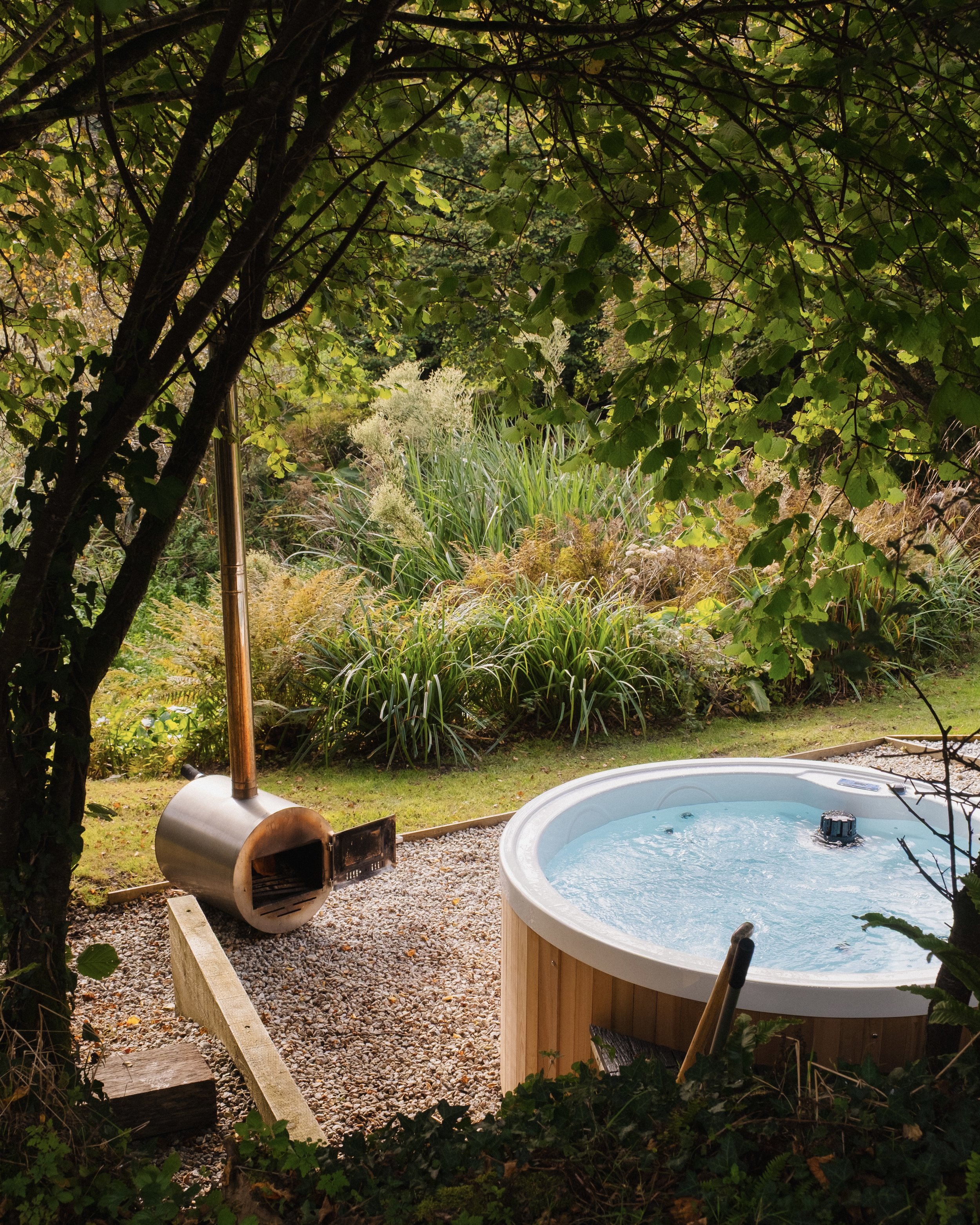 A backyard scene with a hot tub and a wood-fired sauna, surrounded by lush green trees and plants.