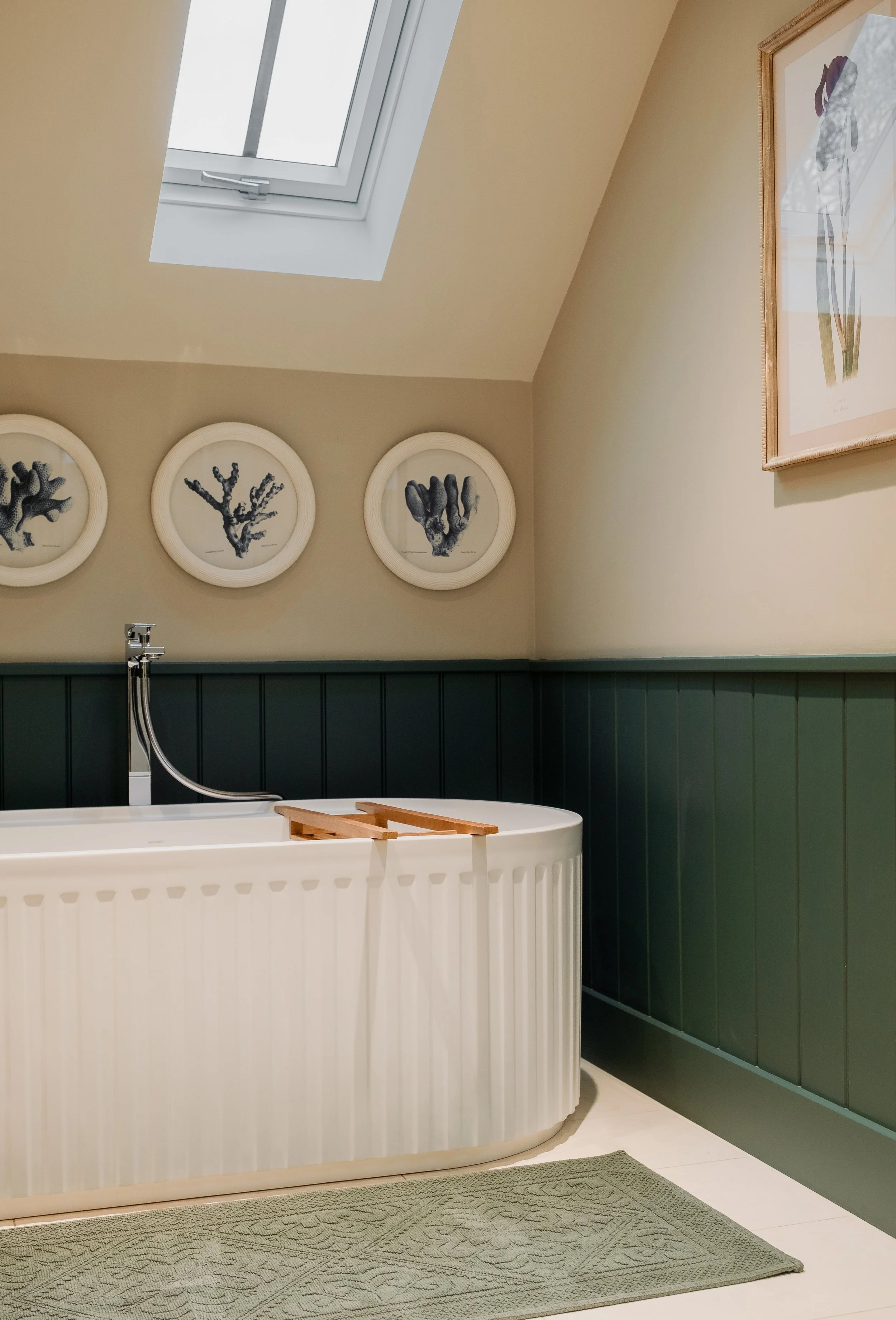 A cozy bathroom with a white bathtub, a skylight window, framed botanical prints on the wall, and a green wainscoting panel.