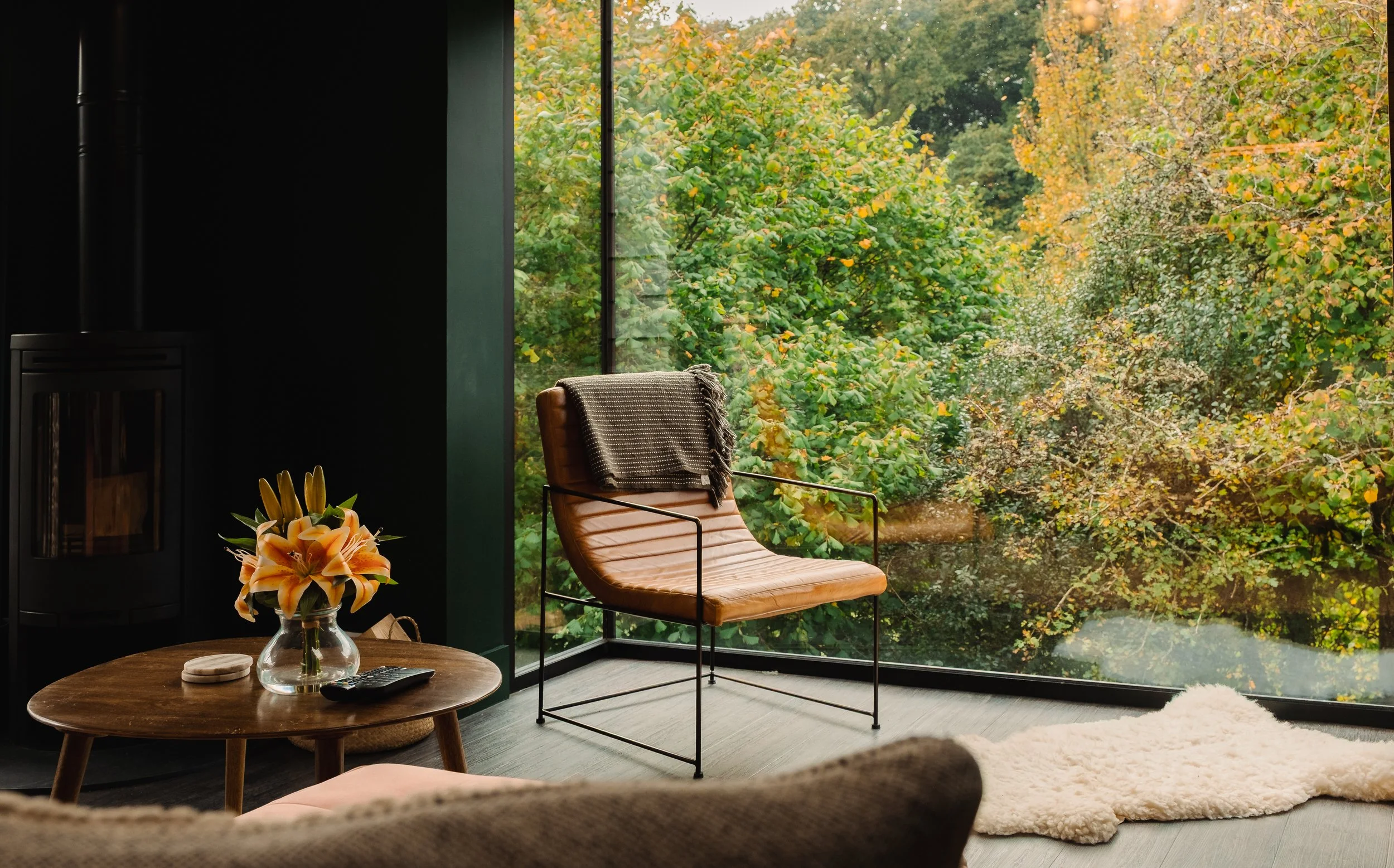 Modern living room with floor-to-ceiling window showcasing trees with green and yellow leaves, a tan leather chair with a gray blanket, a wooden coffee table with a vase of orange lilies, and a cozy cream rug.