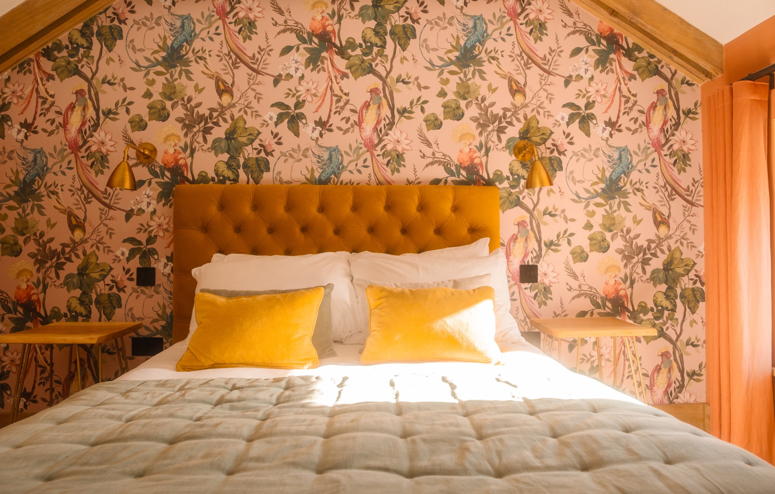 A bedroom featuring a floral wallpaper with birds, a mustard-colored tufted headboard, two yellow pillows, white bedding, two wall-mounted reading lamps, and yellow curtains.