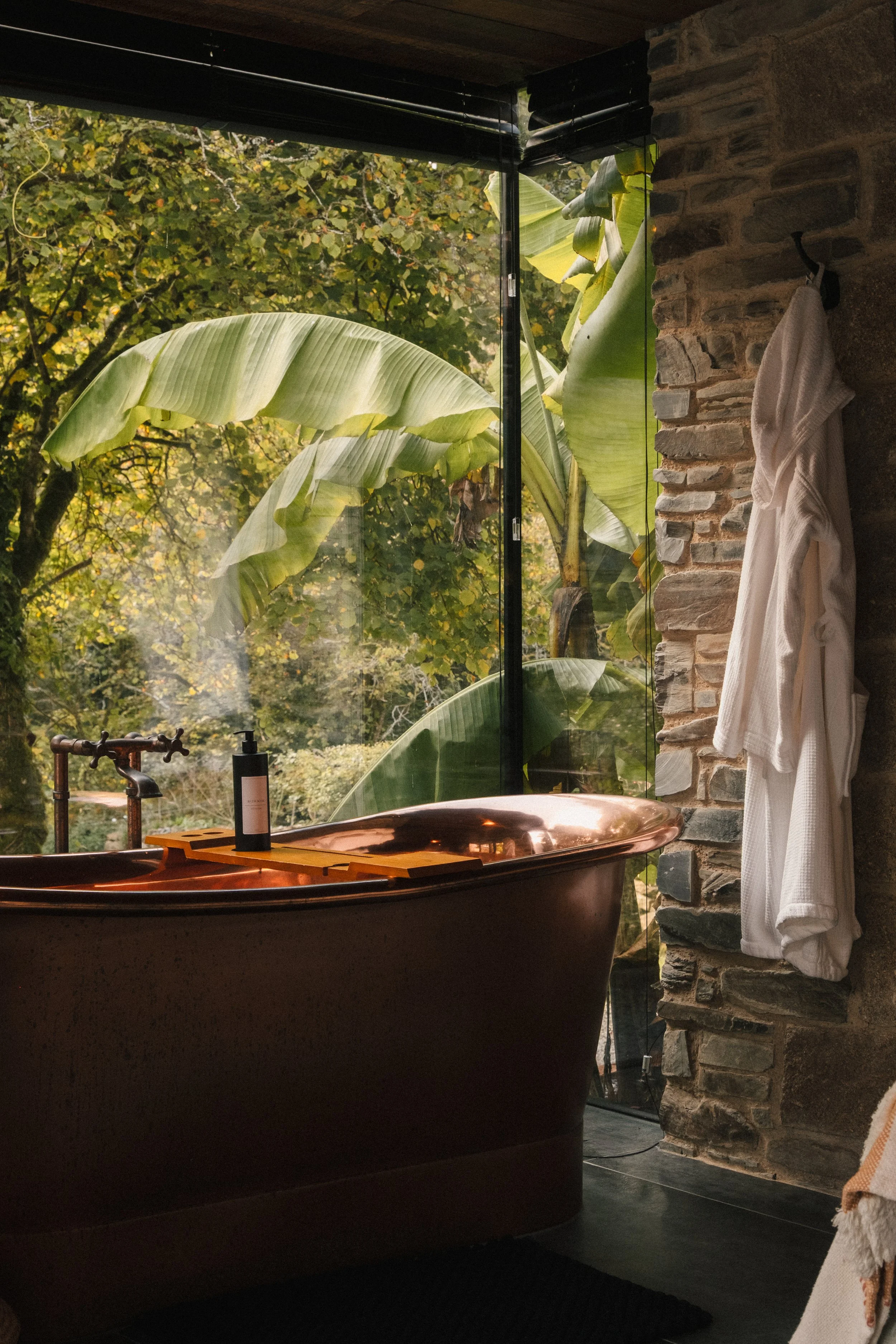 Copper bathtub in a rustic bathroom with a large window showing green leaves outside, a black bottle on the tub's edge, a stone wall, and white towels hanging on hooks.