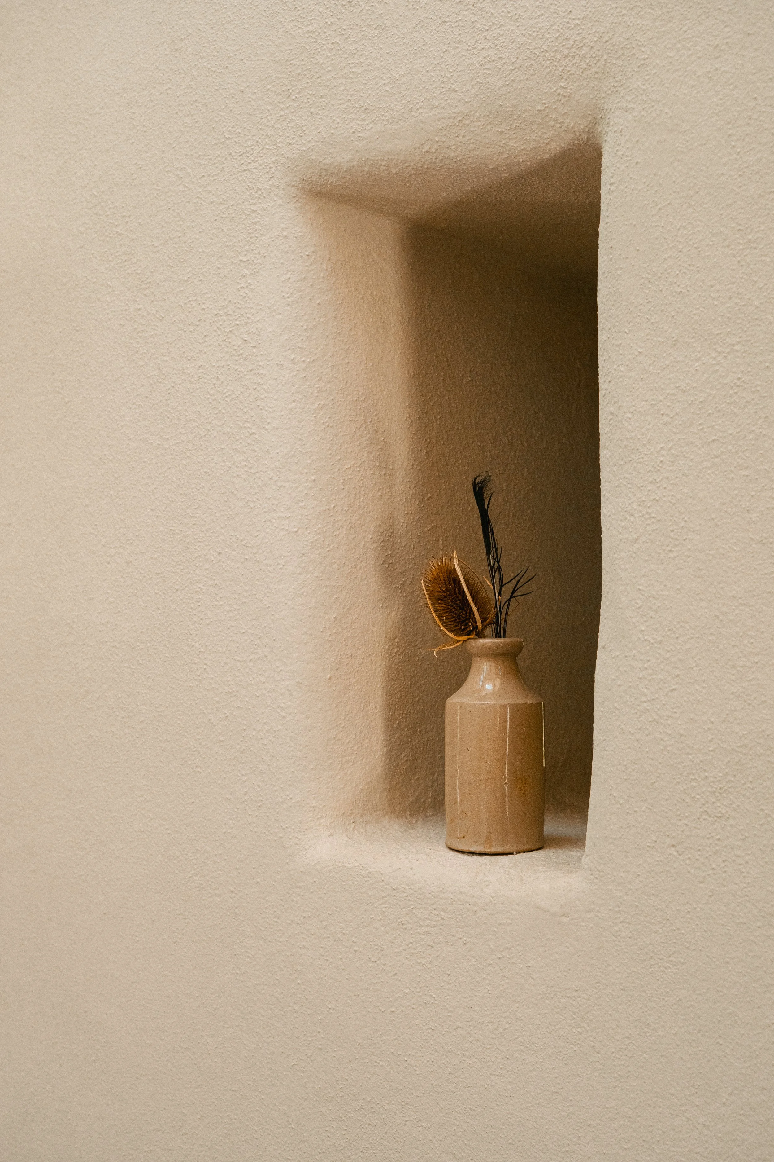 A beige ceramic vase with dried leaves and feathers, placed in a recessed wall nook with textured beige walls.