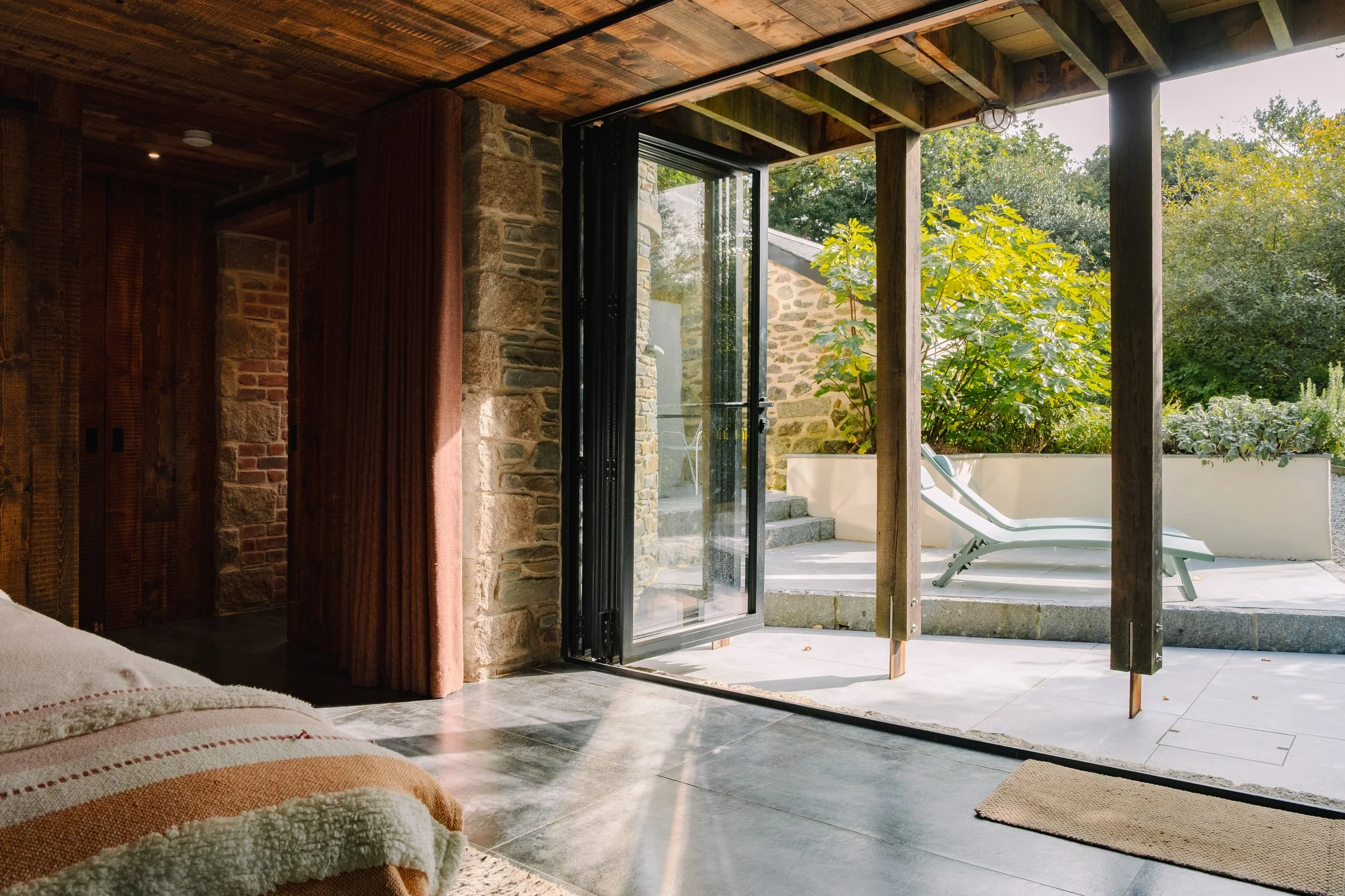 Interior view from a bedroom looking out onto a patio with a lounge chair, greenery, and trees outside.