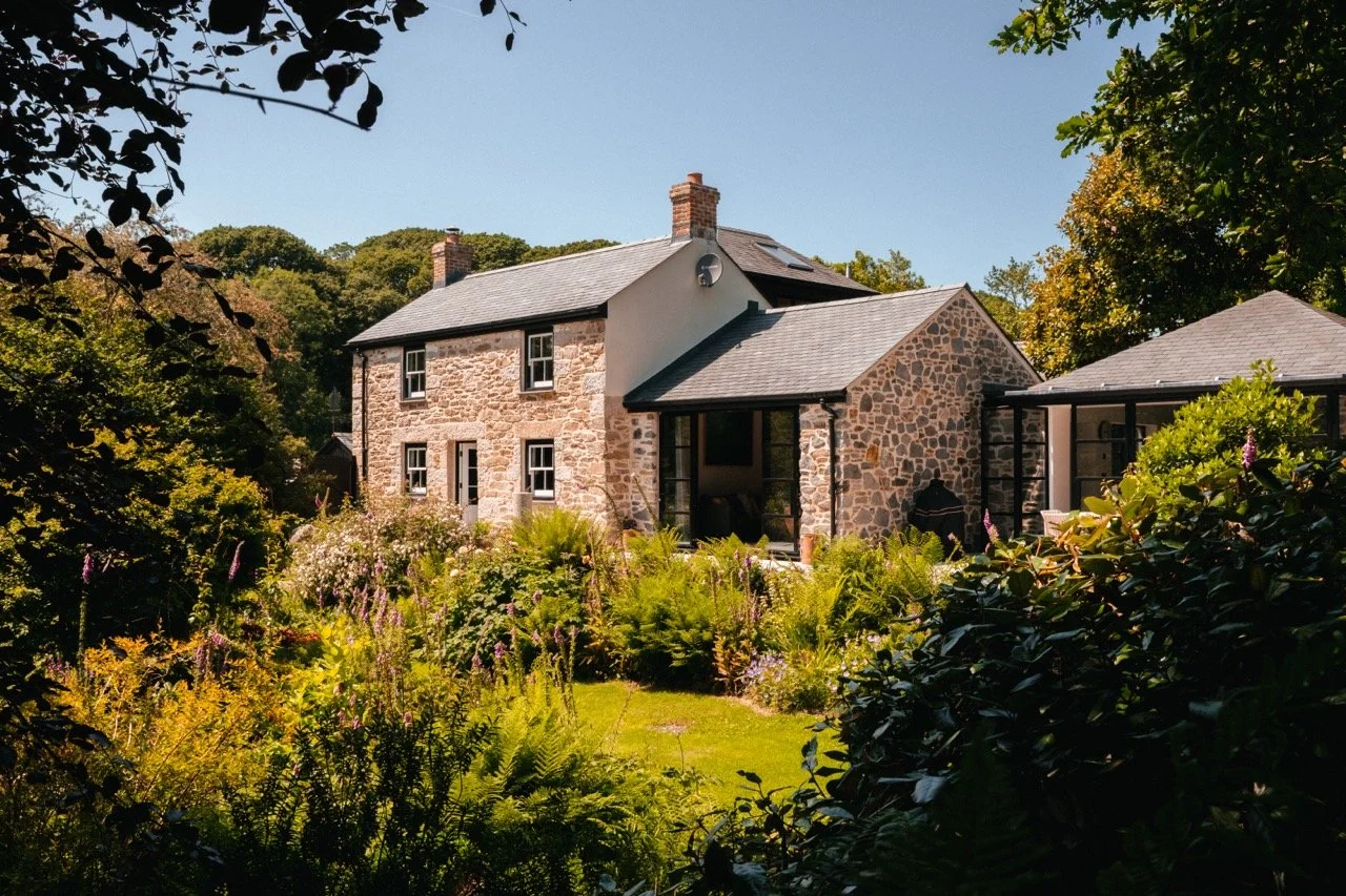 A stone house with a gray shingle roof and surrounded by lush green plants and trees, viewed through a garden with colorful flowers.