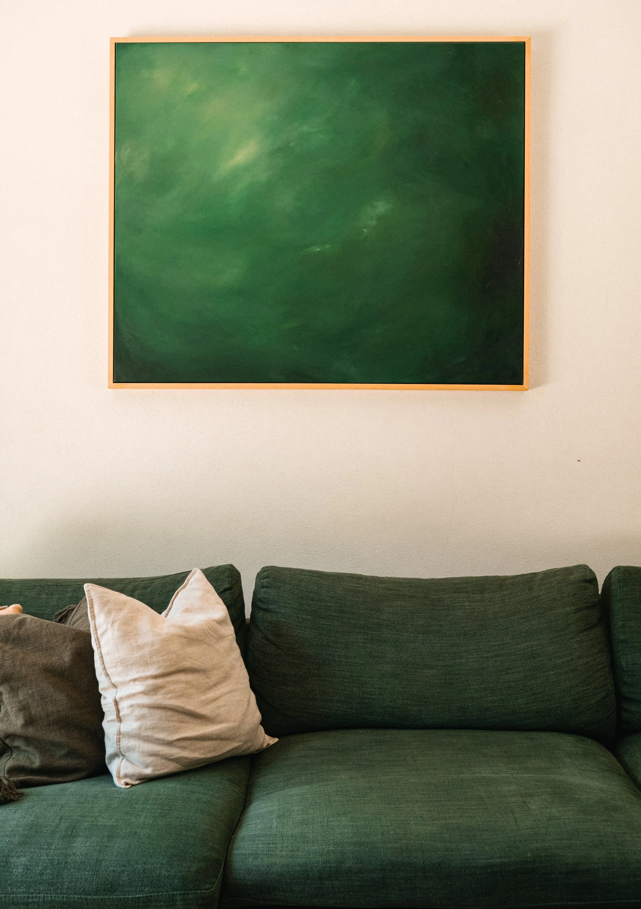 A green abstract painting hung on a plain white wall above a dark green couch with three cushions in shades of beige and gray.