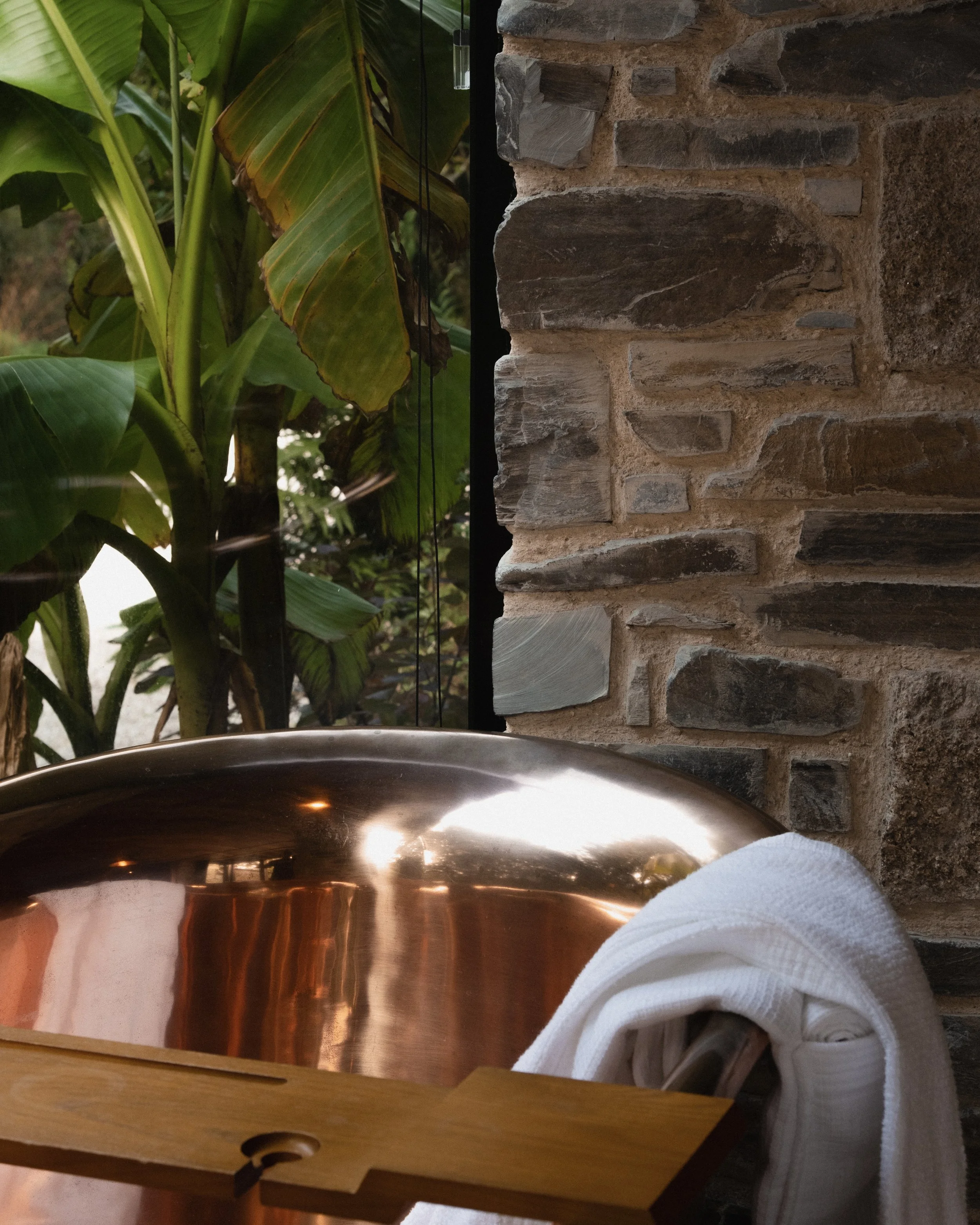 Indoor scene with a copper soaking tub, brick wall, and large green leafy plants outside the window.