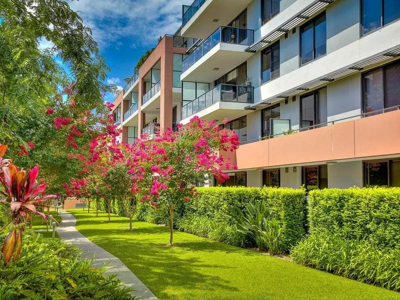 Modern apartment building with balconies, surrounded by lush green trees, pink flowering trees, and a well-maintained lawn on a sunny day.