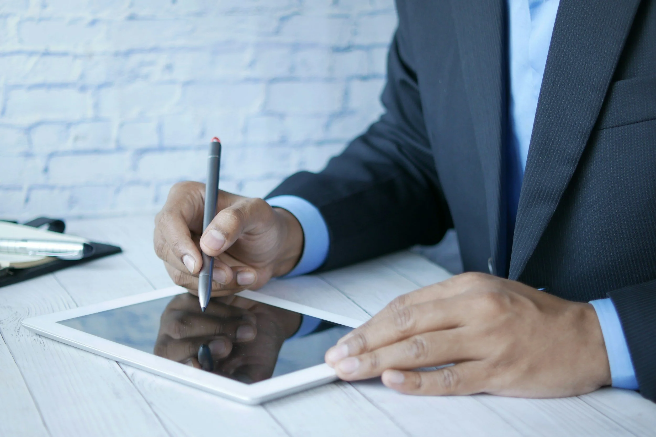 A man in a business suit is sitting at a wooden desk, holding a stylus and interacting with a tablet device.