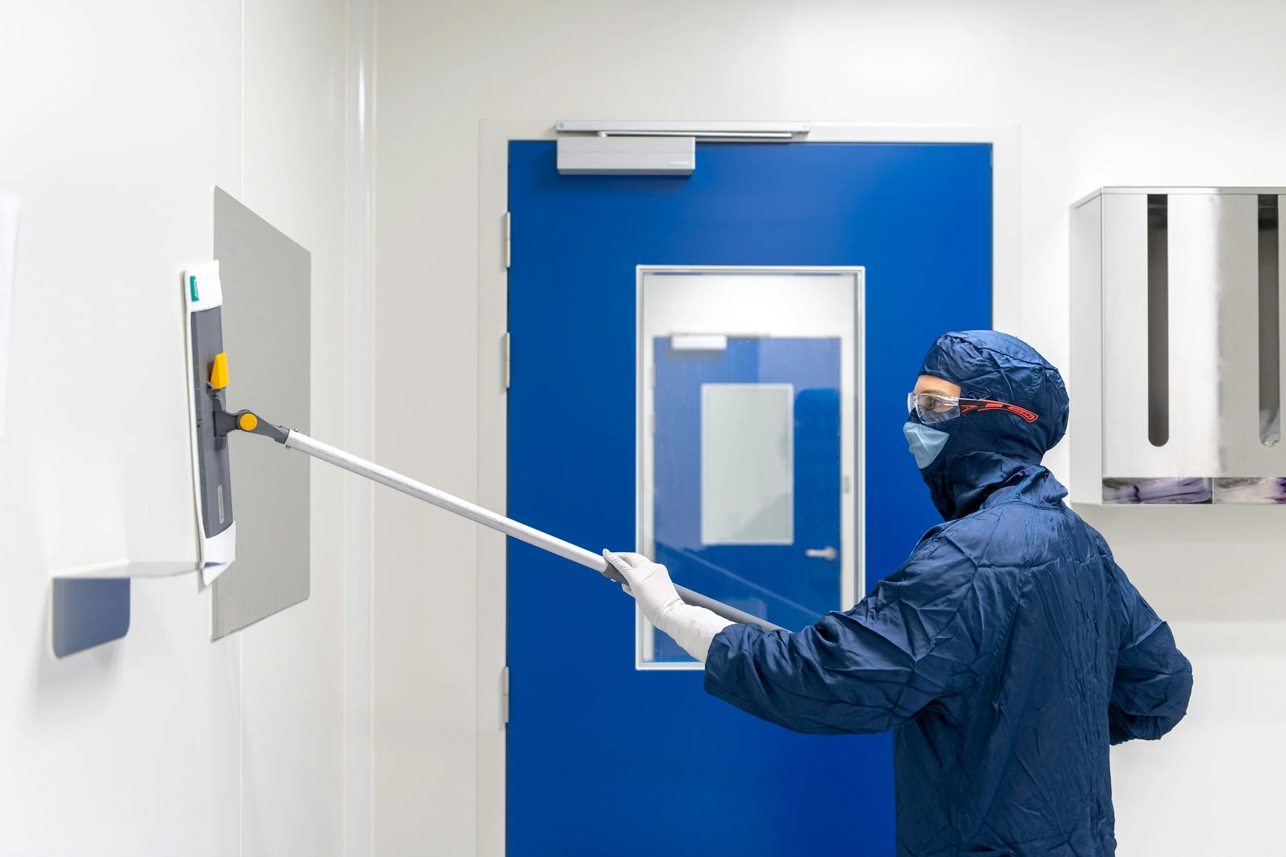 A person in protective gear, including gloves, gown, goggles, and mask, disinfects a wall with a long-handled sprayer in a clinical setting.