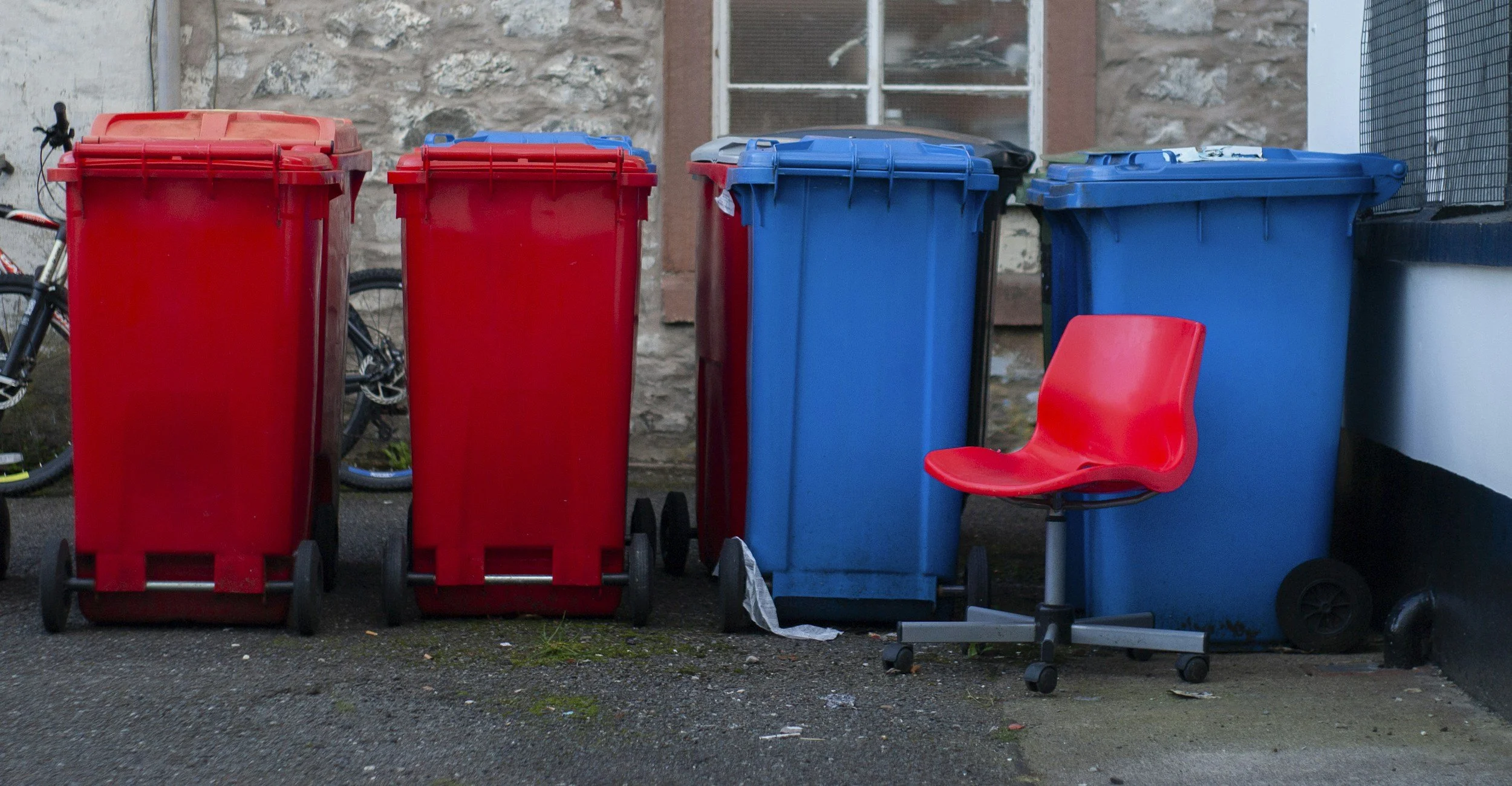 Three trash bins, two red and one blue, with a red rolling office chair placed in front of them on a concrete surface.