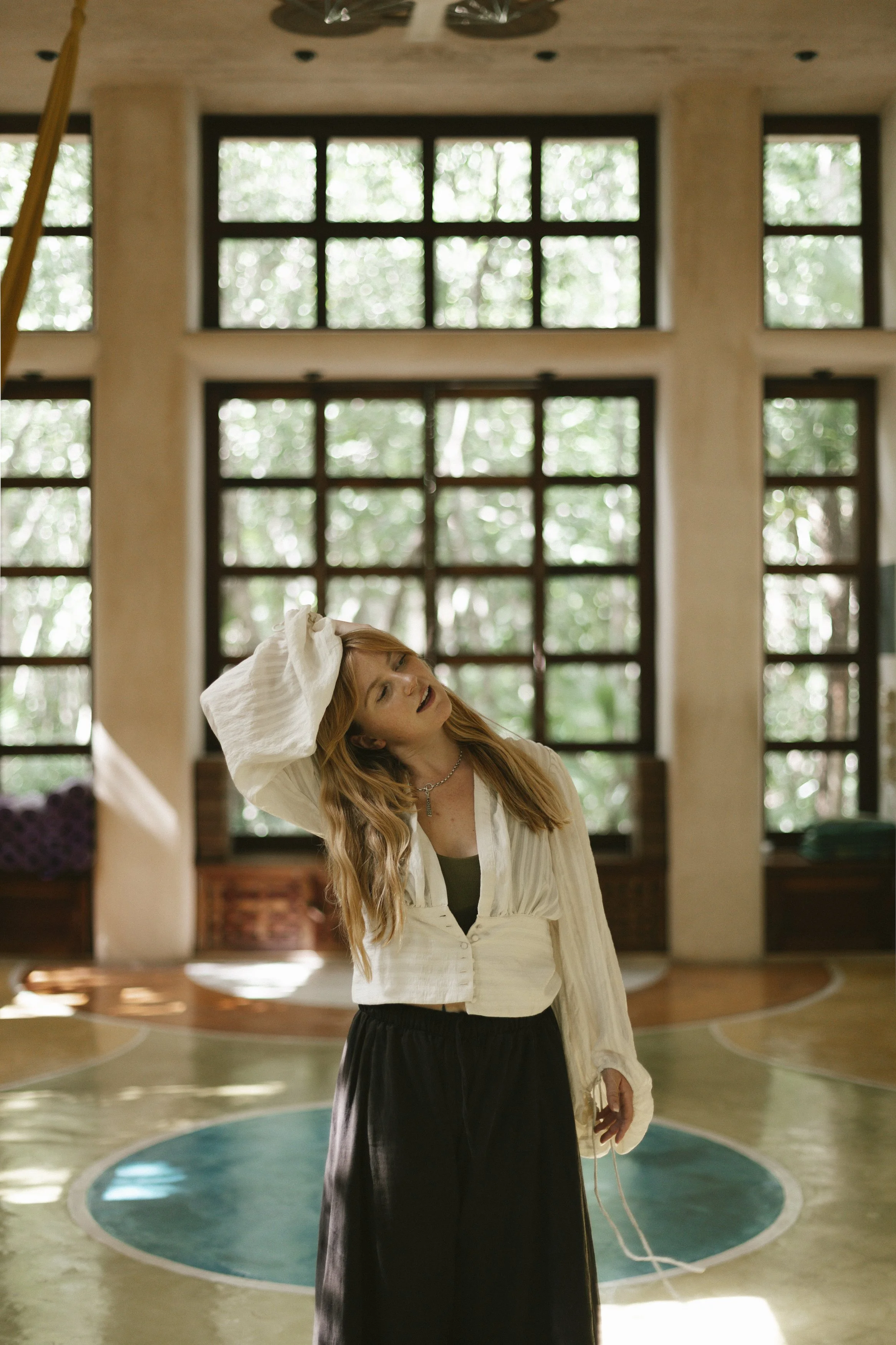 a vinyasa yoga teacher in Tulum with ginger hair and a flowy white top with long sleeves, taken in holistika with incredible light and design of the yoga studio