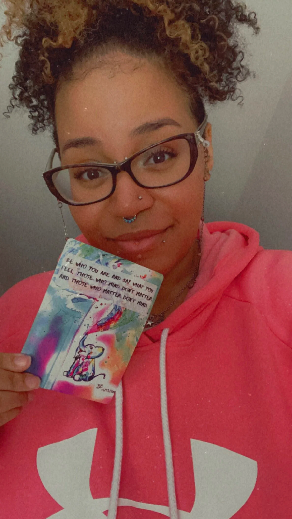 A young woman with curly hair, glasses, and facial piercings holding a colorful card with a rainbow, an elephant, and an uplifting quote in front of a plain wall, wearing a pink hoodie with a white logo.