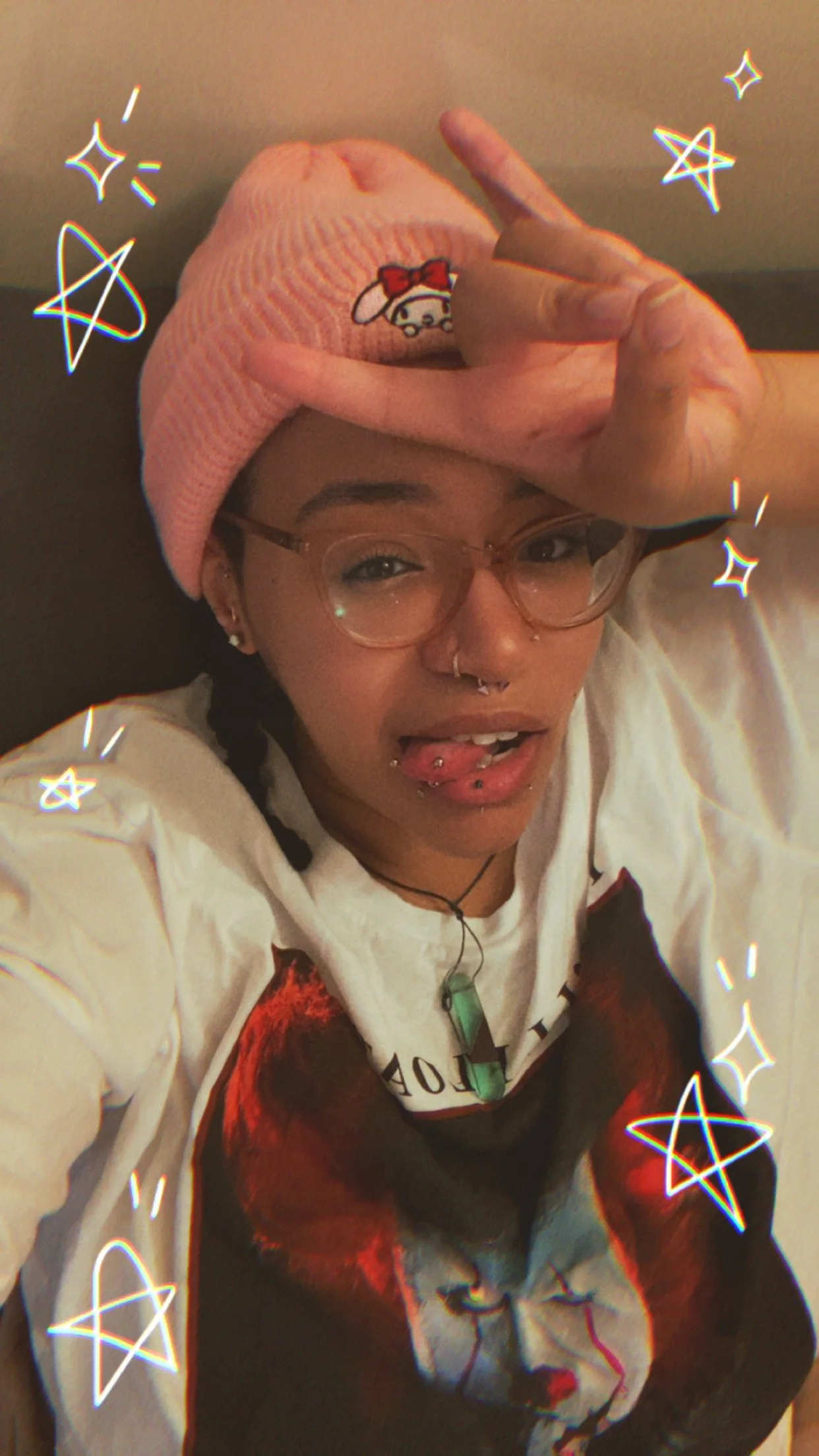 A young woman with glasses, piercings, and tattoos wearing a pink Hello Kitty beanie, a white shirt with a graphic print, and a necklace, making a peace sign with her hand on her forehead. She is sticking out her tongue with piercings. The photo has stickers of stars and sparkles.