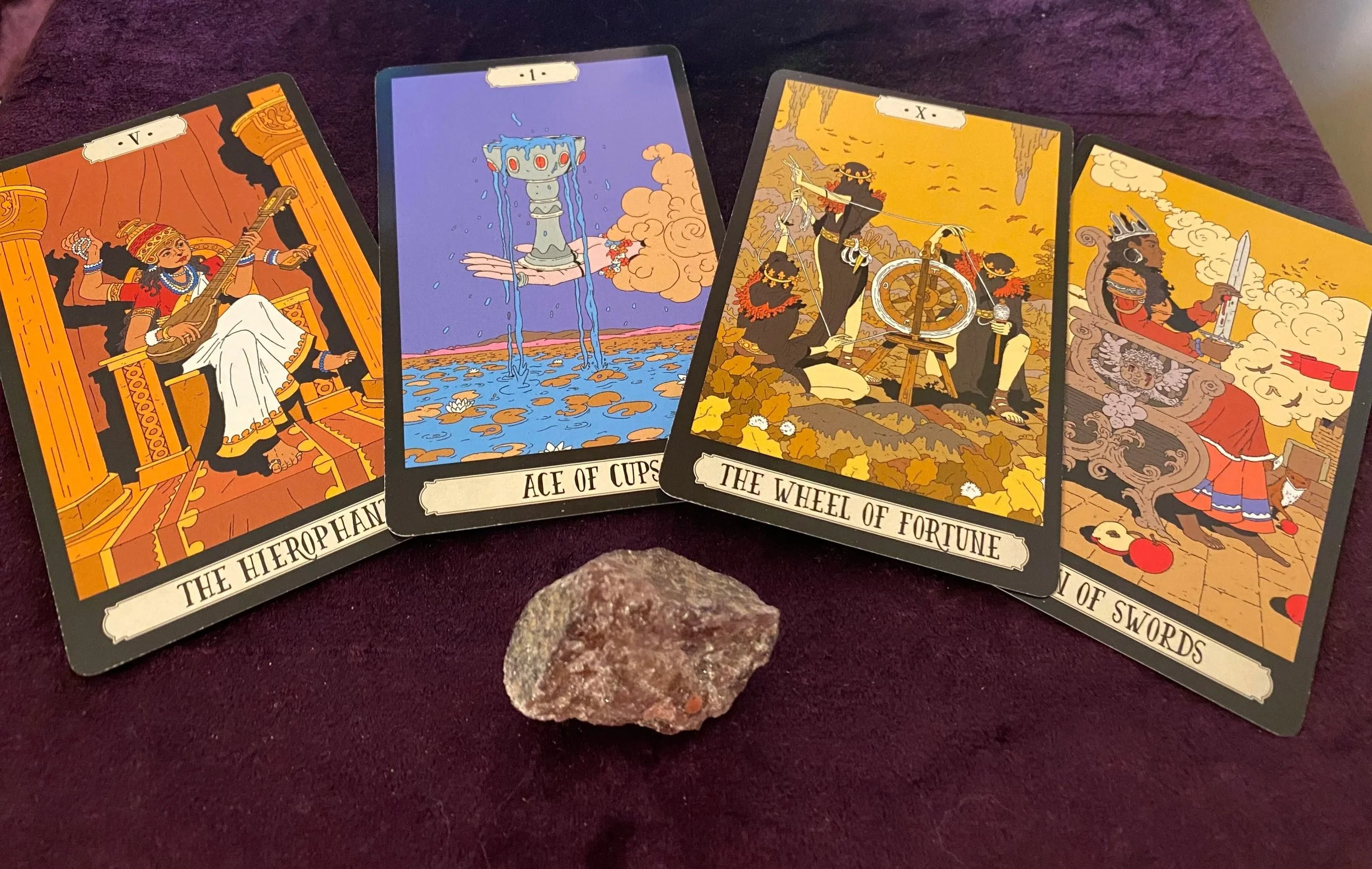 Four tarot cards laid out on a dark red cloth with a small rock in front of them. The cards are labeled 'The Hierophant', 'Ace of Cups', 'The Wheel of Fortune', and 'Knight of Swords', each featuring colorful illustrations and symbols.
