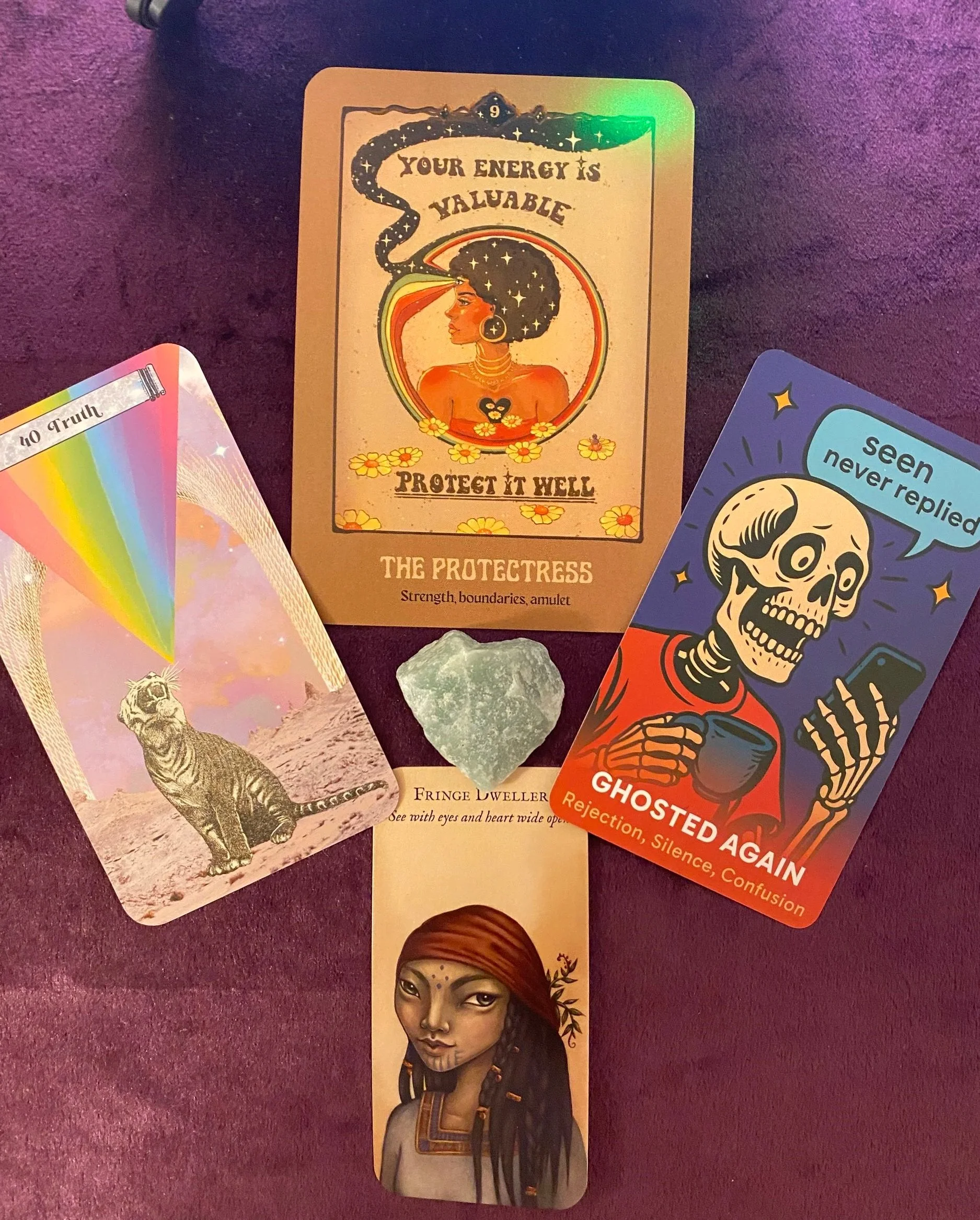 A collection of four cards and a green stone on a purple surface. The central card reads 'Your energy is valuable, protect it well' and shows a woman with natural hair and a black snake. The card below it is a portrait of a woman with dark skin and long dreadlocks. To the right, a skeleton character holding a cup and a phone says, 'Seen never replied,' with the caption 'Ghosted Again' and words 'rejection, silence, confusion.' To the left, a colorful card shows a tiger in a desert with a rainbow-shaped element and some text at the top.