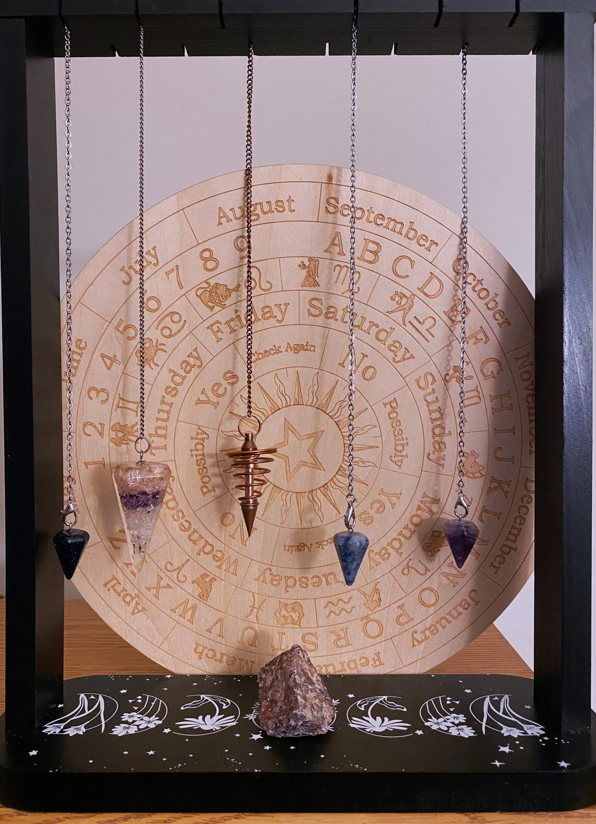 A display with a circular wooden zodiac calendar in the background, four necklaces with large gemstone pendants hanging from a frame, and a rough gemstone stone on the platform.