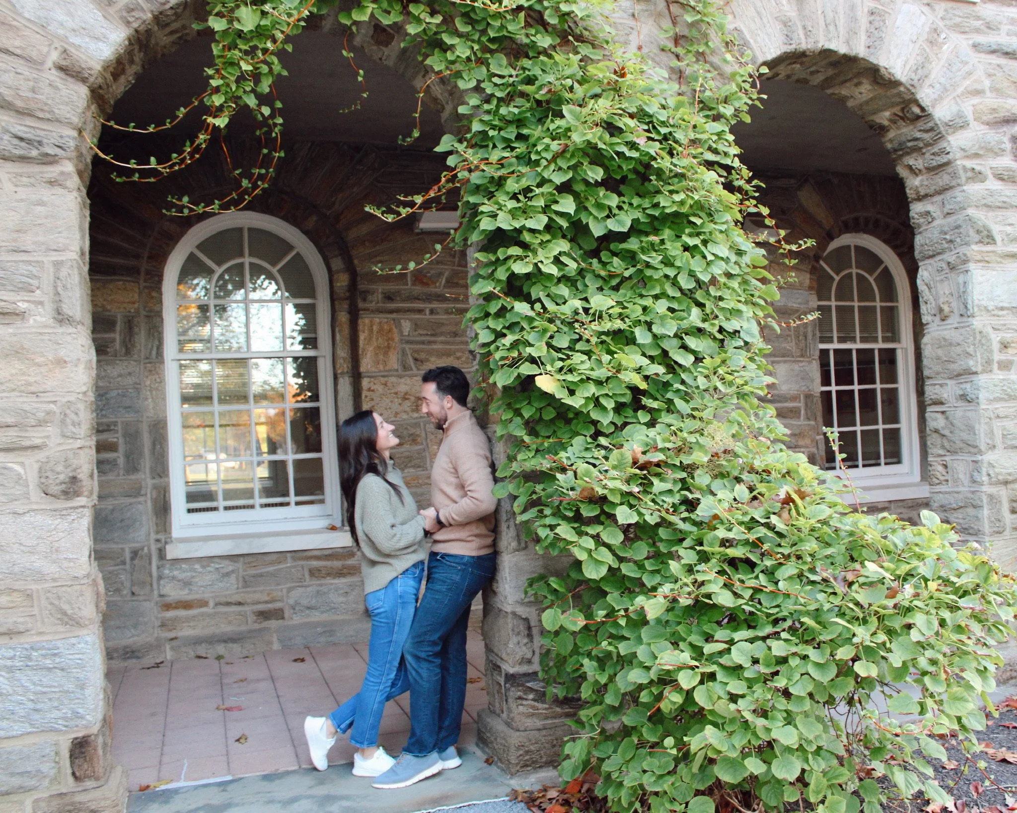 A couple standing close together, smiling and holding hands outside a stone house with ivy-covered columns and large arched windows.