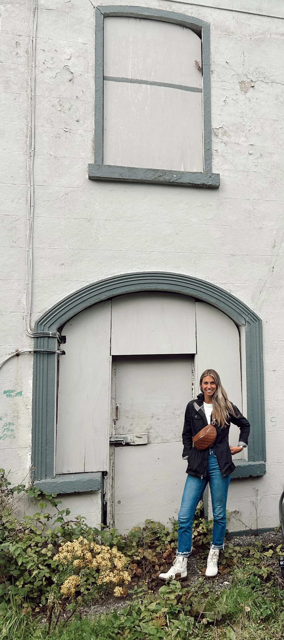 A woman with long blonde hair smiling, standing outdoors in front of a white, weathered building with boarded-up windows and door, wearing white roller skates, blue jeans, and a black jacket.