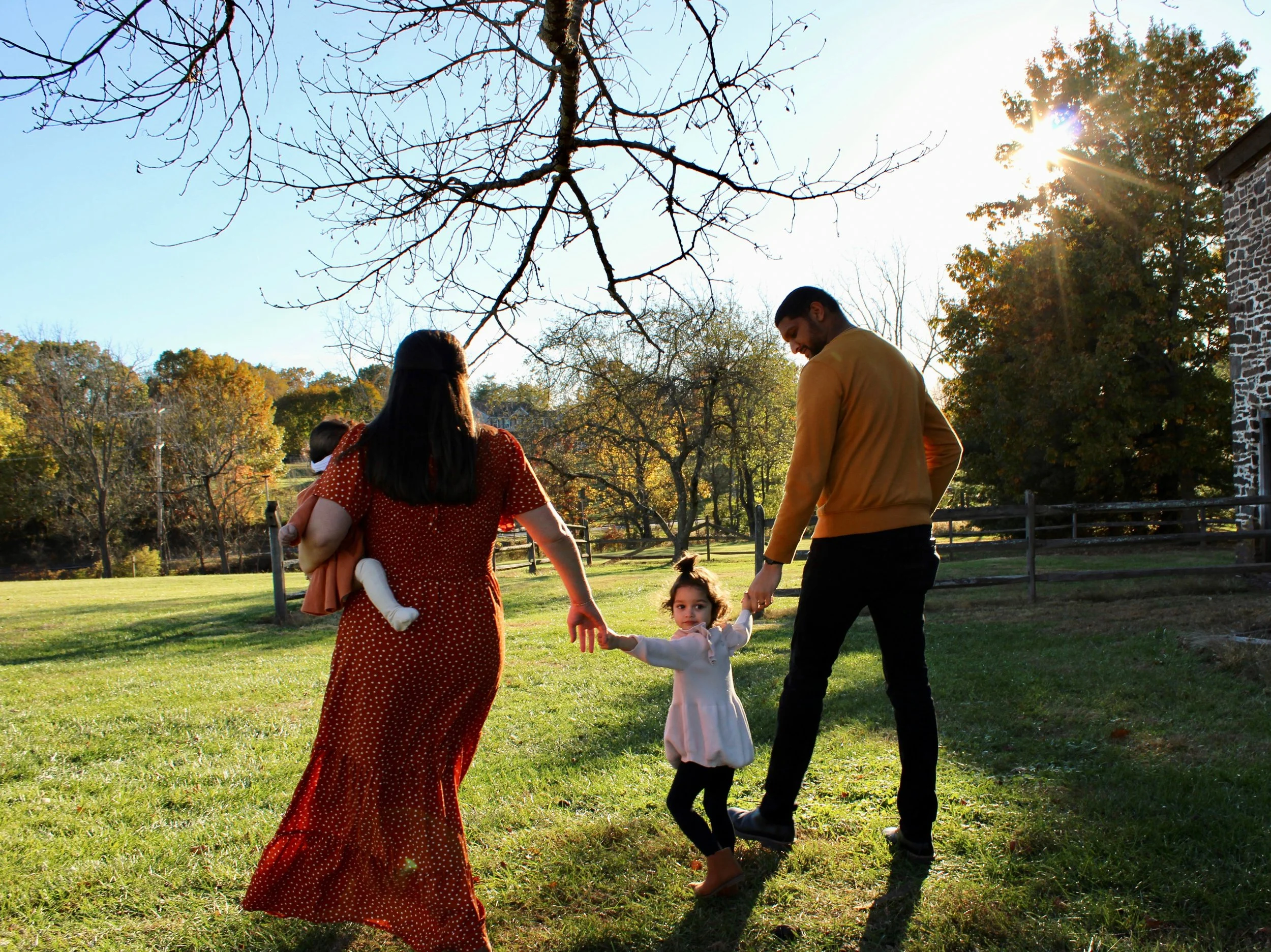 Family of four, including a woman holding a small child, a man, and a young girl, playing and holding hands in a grassy park on a sunny day.