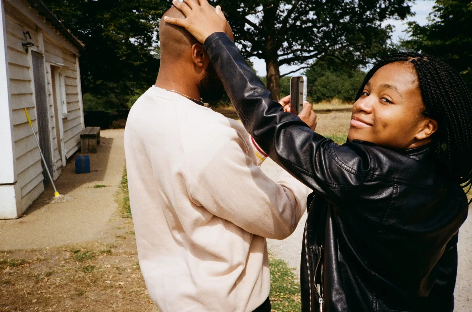 A young woman smiling while playfully patting a young man's bald head, as he looks at his phone outdoors near a white wooden building and trees.