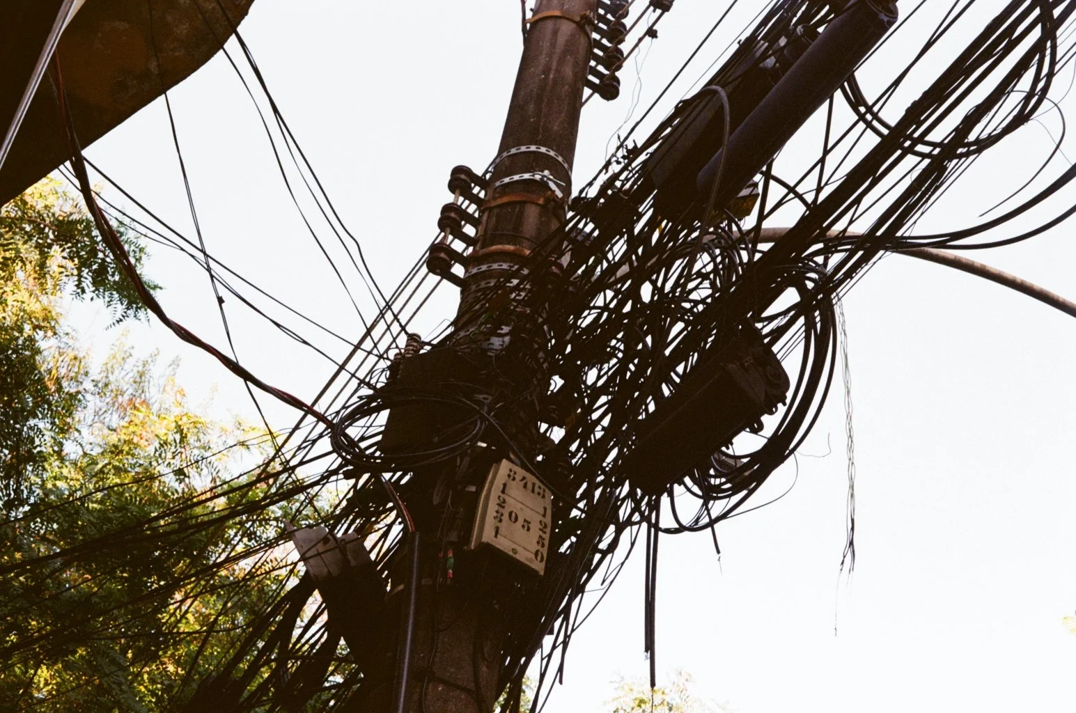Close-up view of a utility pole with an extensive mass of tangled wires and cables, with a background of the sky and some tree branches.