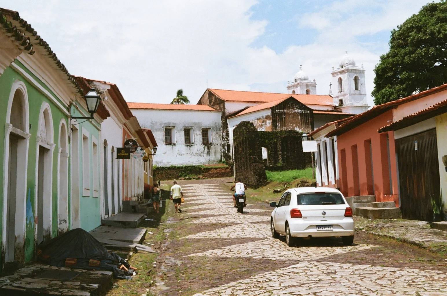 A cobblestone street in a historic town with colorful buildings, two church towers in the background, a white car parked on the right, and people walking along the street.