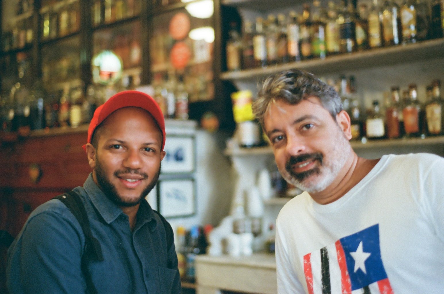 Two men smiling and looking at the camera in a cafe or bar setting with shelves of jars and bottles in the background.