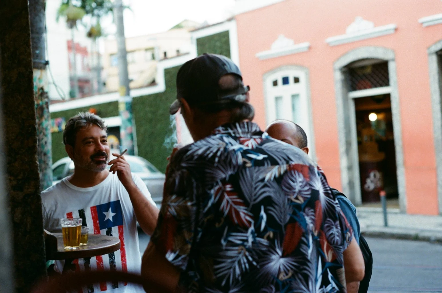 Group of men socializing on city street in front of colorful buildings, one wearing a white t-shirt with a flag design, two others wearing patterned shirts, one with a cap, at dusk.
