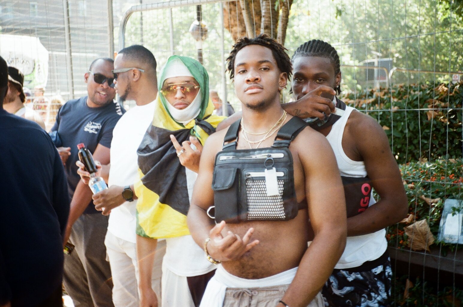 A group of young people standing in line outdoors, with one person in the foreground making a hand gesture. The group is casually dressed, and some are wearing accessories like sunglasses and chains. There is greenery and a wire fence in the backgrou