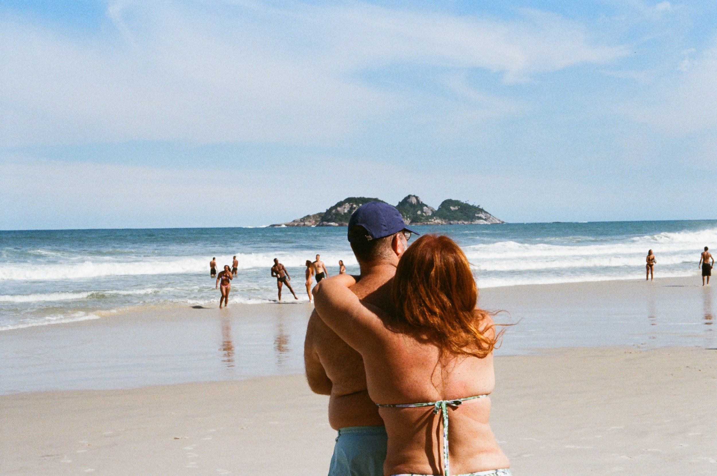 Couple embracing on a beach with people wading in the ocean, and rocky islands in the background under a partly cloudy sky.