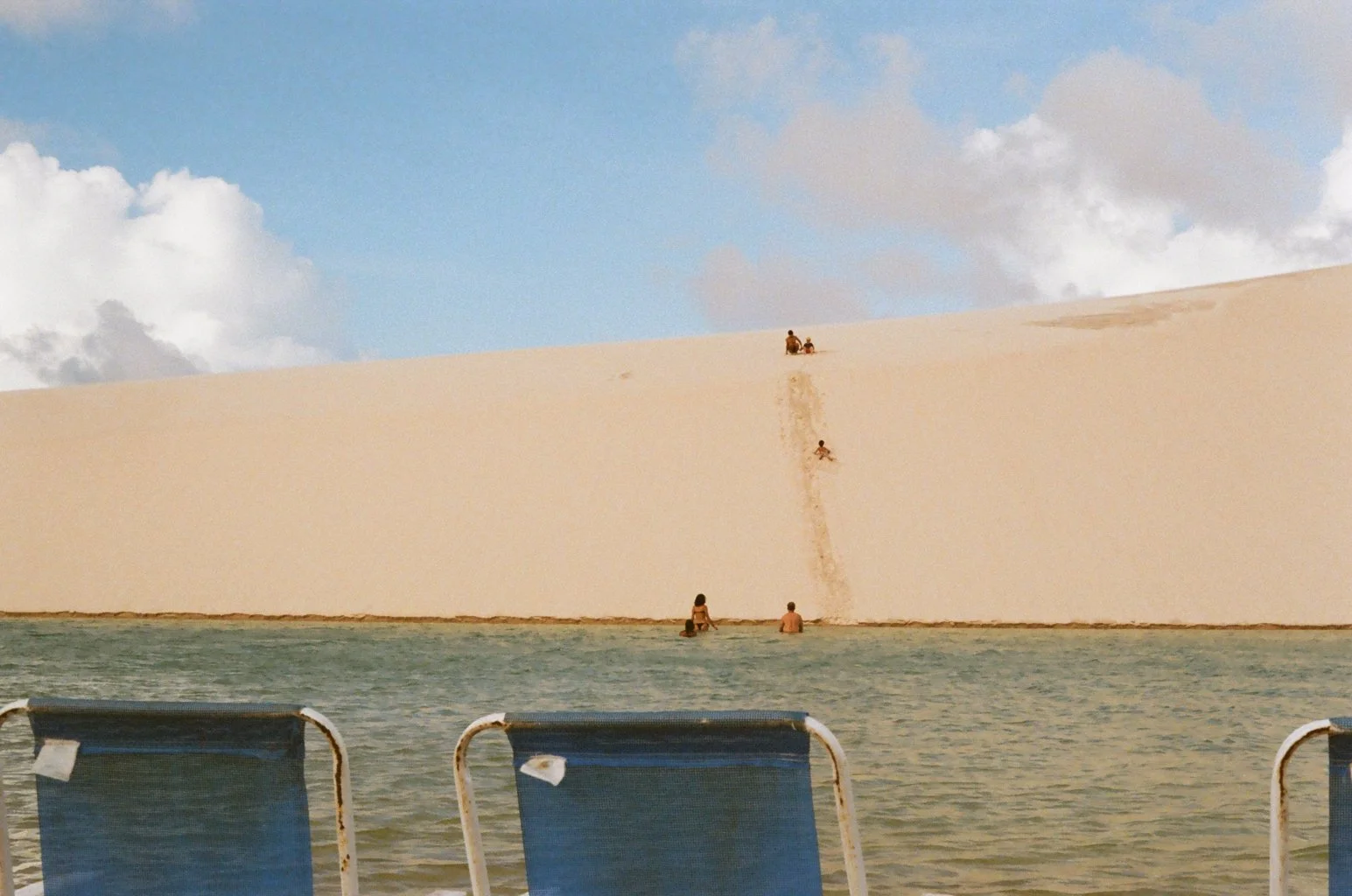 A sandy desert with a small group of people climbing a large sand dune, some sitting at the bottom near water, under a partly cloudy sky.