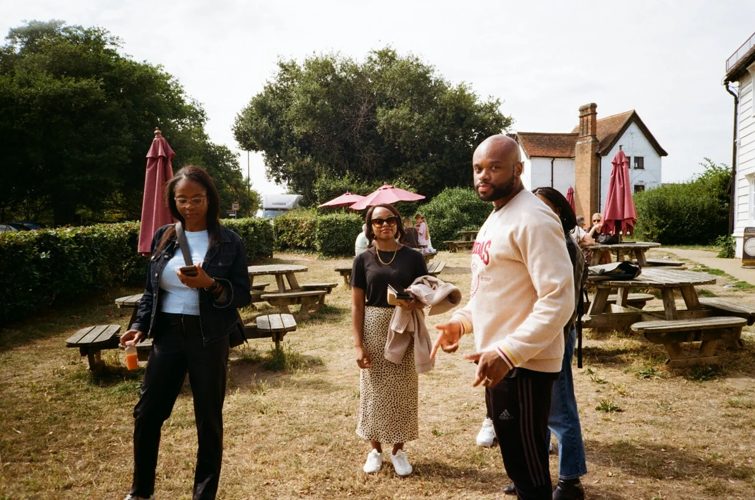 Group of four Black adults outdoors at a park with wooden picnic tables, some holding phones, surrounded by trees and buildings.